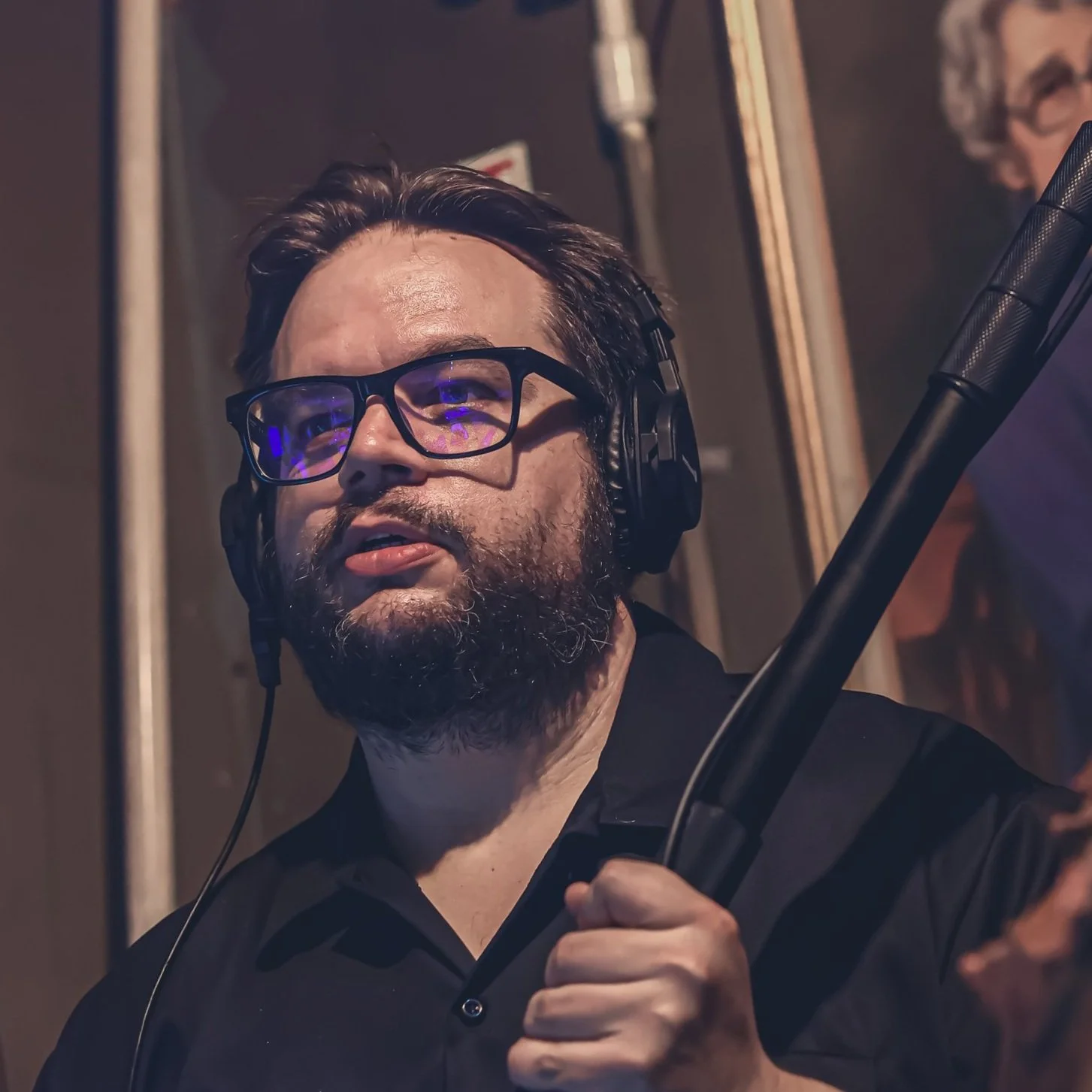 A man with glasses and a beard in a recording studio, wearing headphones and holding a boom microphone.