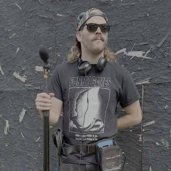 Man in black T-shirt with graphic design, sunglasses, cap, holding a boom microphone, standing against a black textured wall.