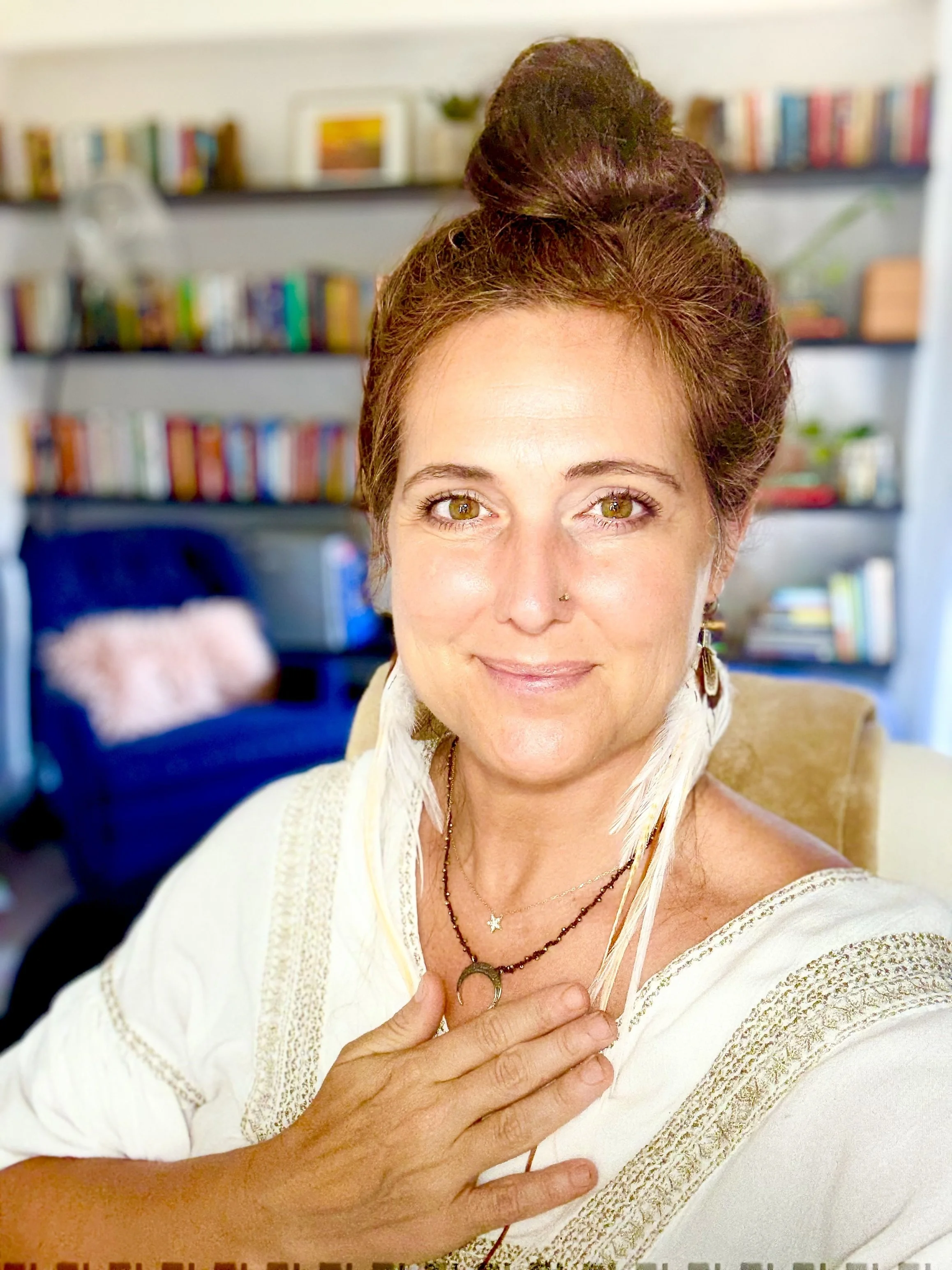 A woman with reddish hair styled in an updo, wearing a white embroidered top and layered necklaces, holding her hand over her chest in a gesture of sincerity or gratitude, with a background of bookshelves filled with colorful books and decor.
