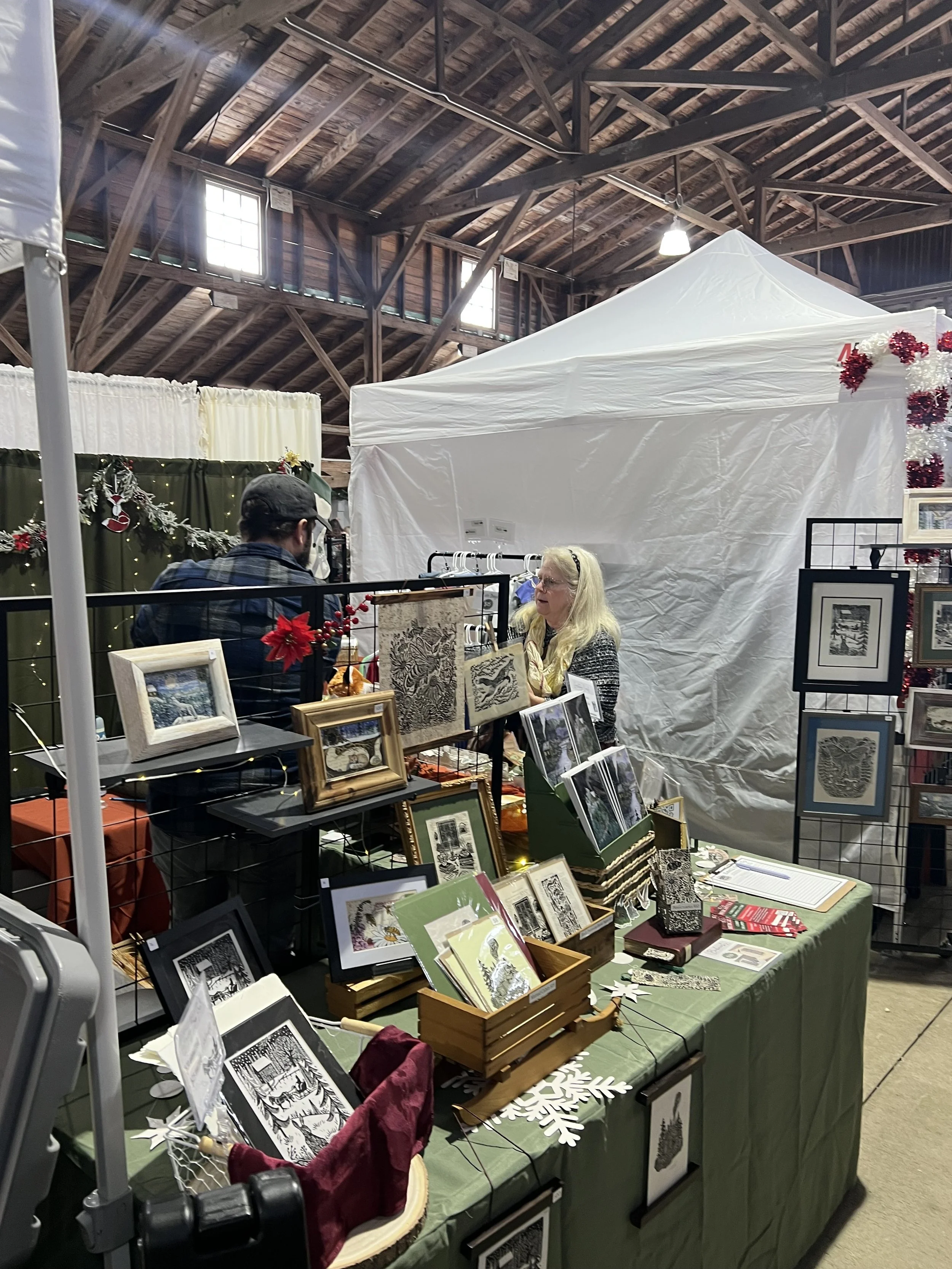 An indoor craft fair or holiday market stall with framed artwork and decorative items, with a woman and a man talking, surrounded by festive decorations, in a wooden-roofed hall with high windows.