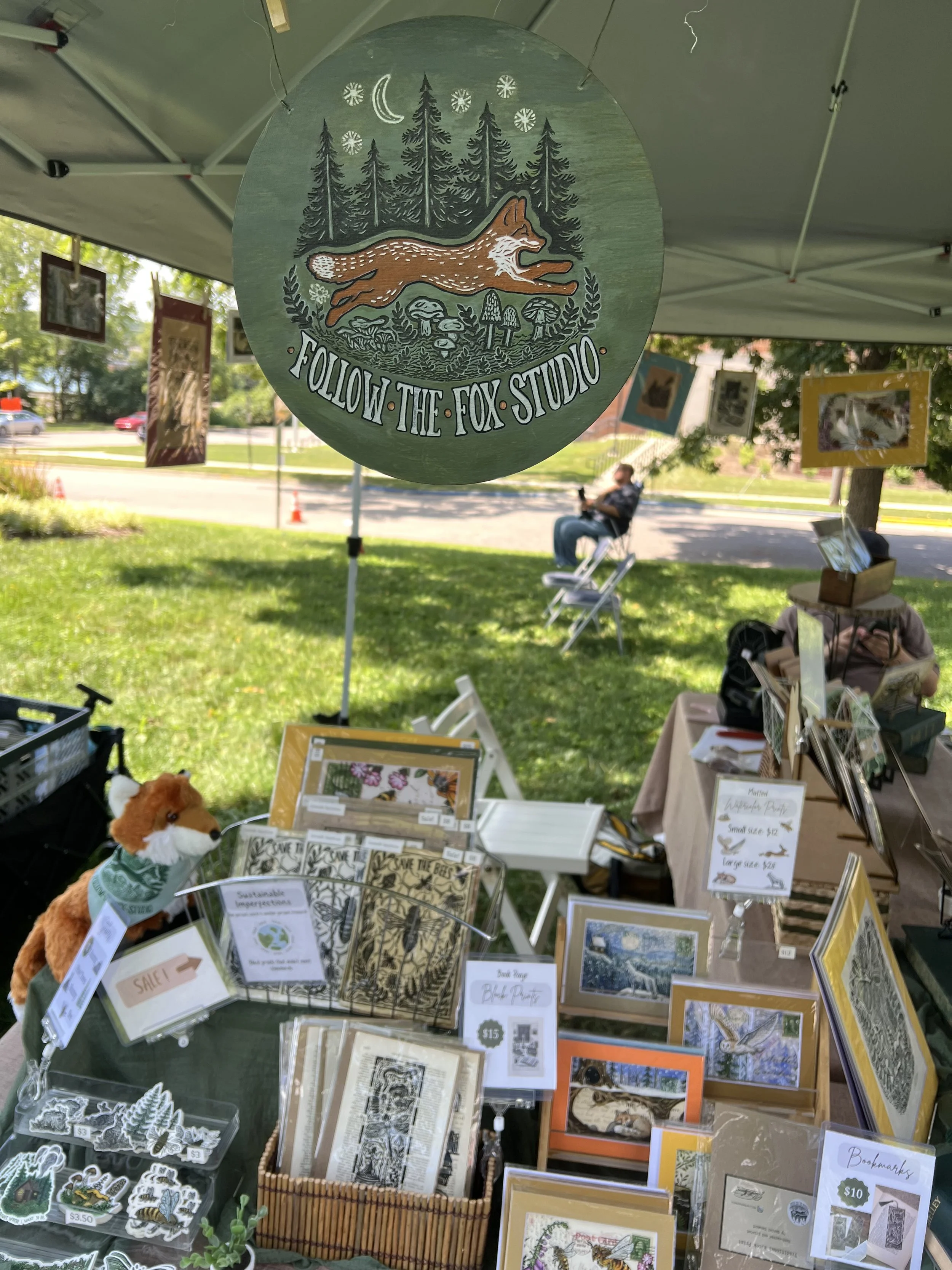 Outdoor art stall with a hanging circular sign that reads 'Follow the Fox Studio' and features a fox running in a forest with trees and stars in the background. The stall displays framed artwork, stickers, and plush toys.