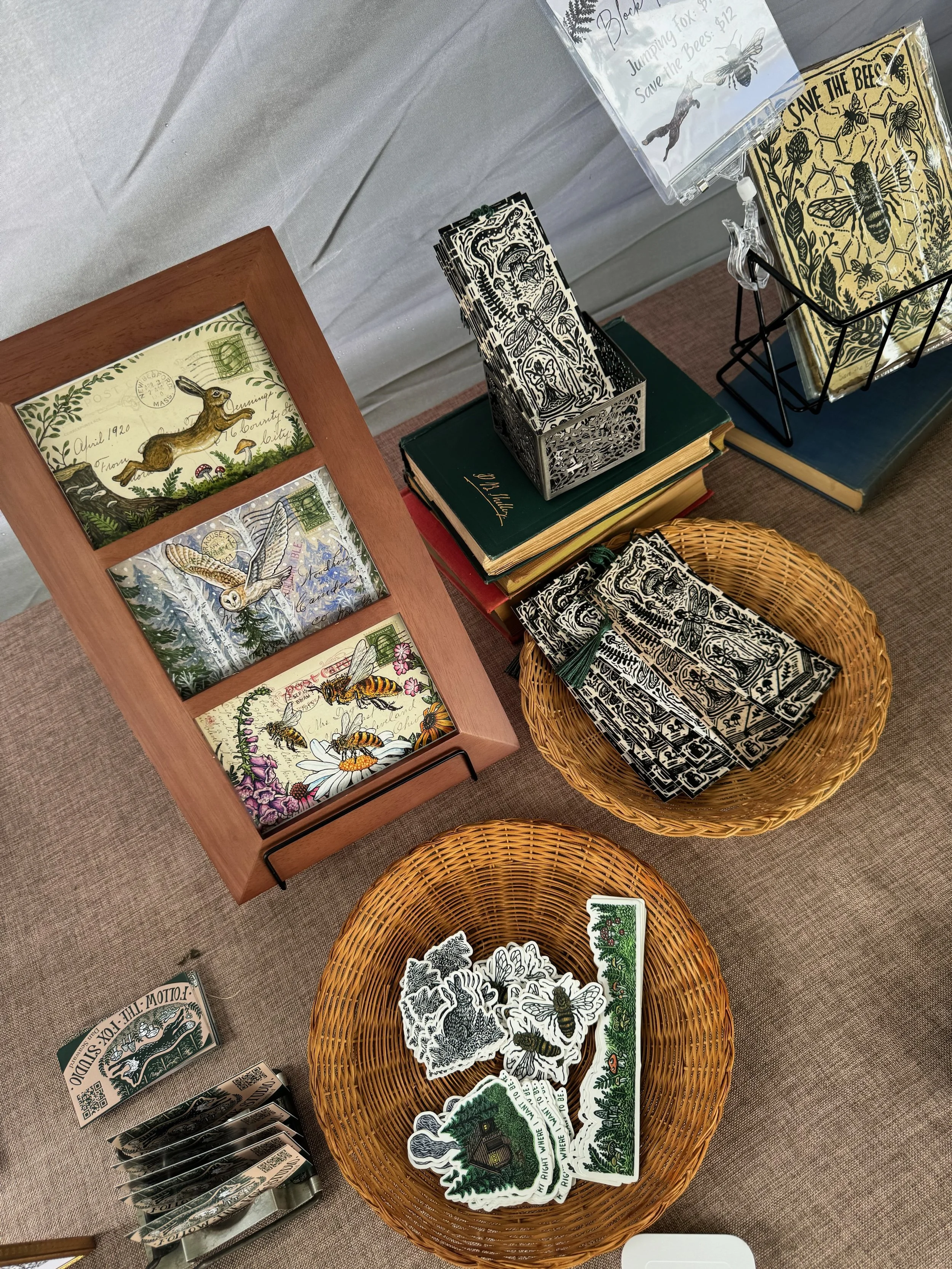 A display of bee-themed stickers, cards, and artwork on a table, with books and a wire basket holding paper items, including a 'Save the Bees' poster and illustrations of bees and flowers.