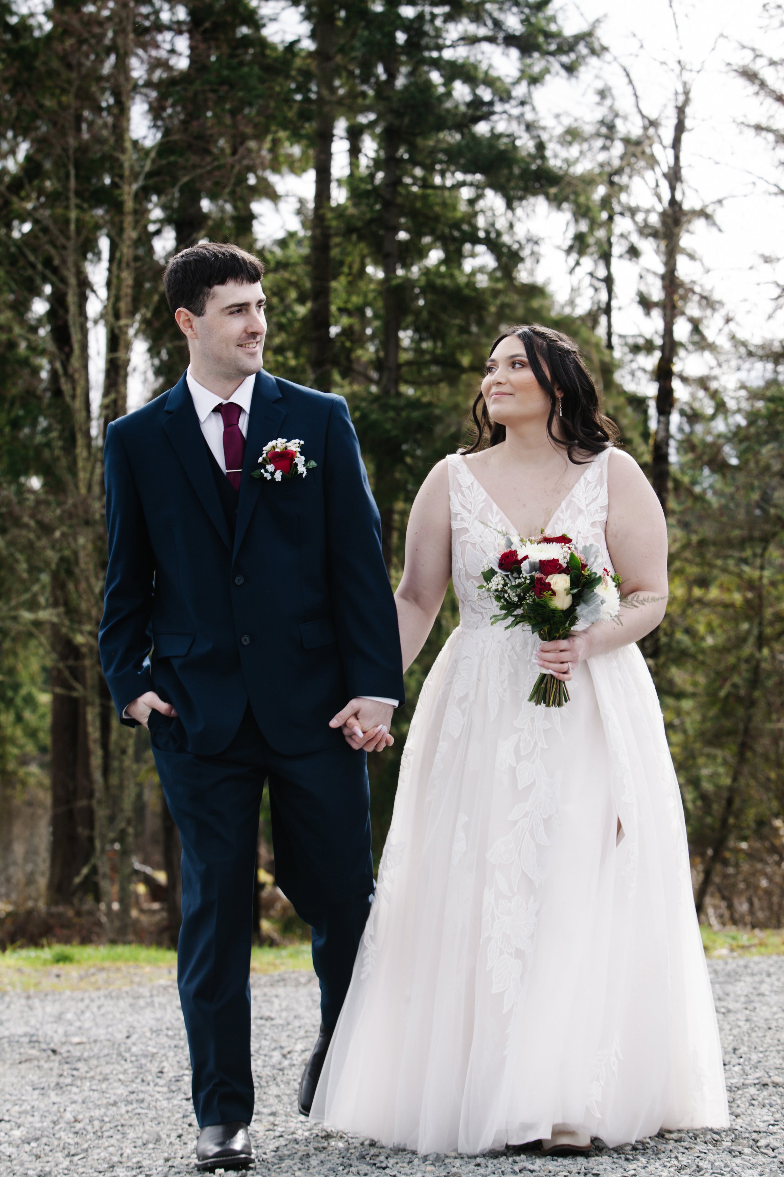 A bride and groom holding hands outdoors in a wooded area with tall trees, dressed in wedding attire. The groom wears a navy suit with a boutonniere, and the bride wears a white gown with lace details, holding a bouquet of red, white, and green flowe