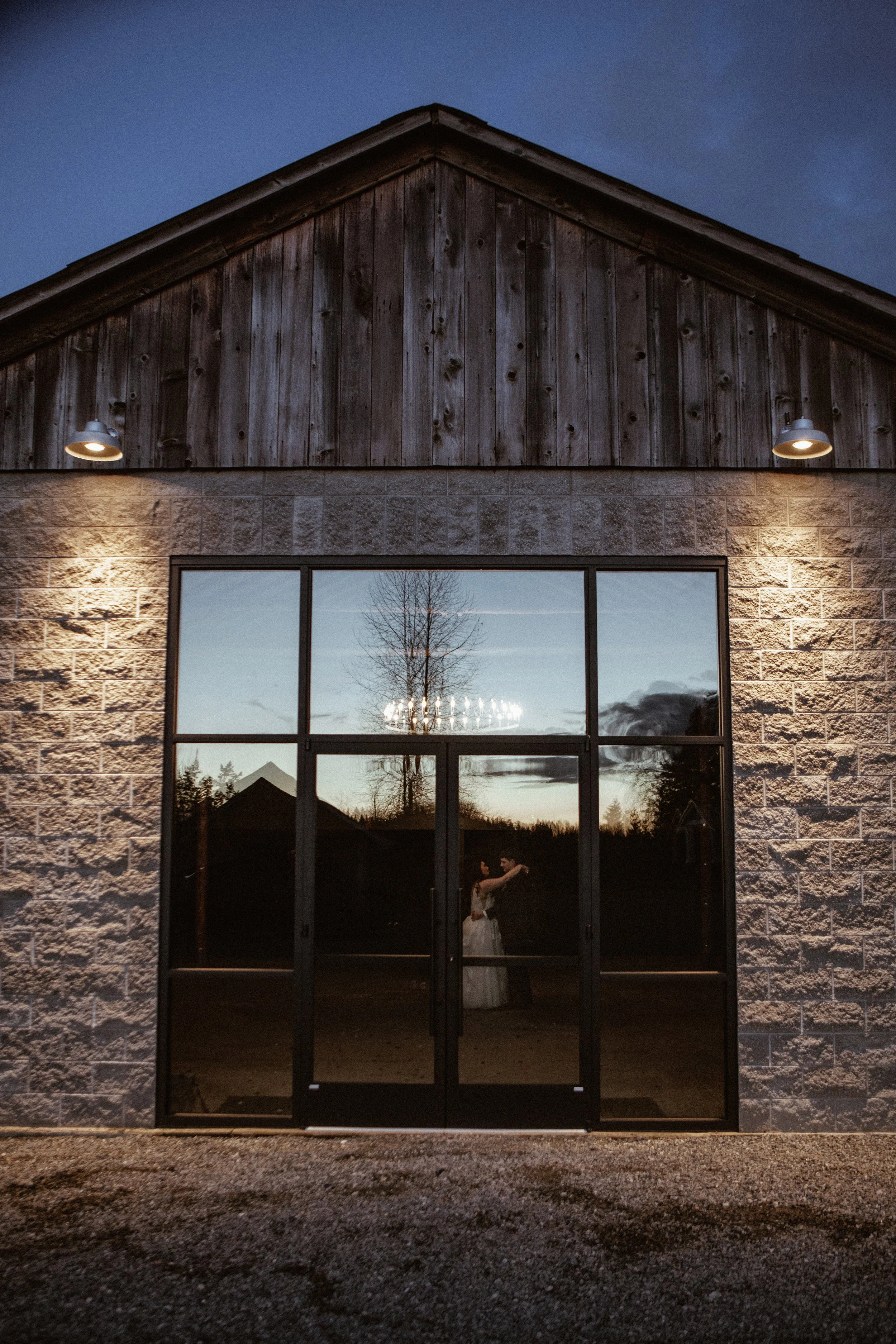 A couple inside a building, seen through large glass windows, suggests they are embracing or dancing. The building has a stone lower wall, wooden upper wall, and is lit by exterior lights. The scene is set during dusk or early evening, with a reflect