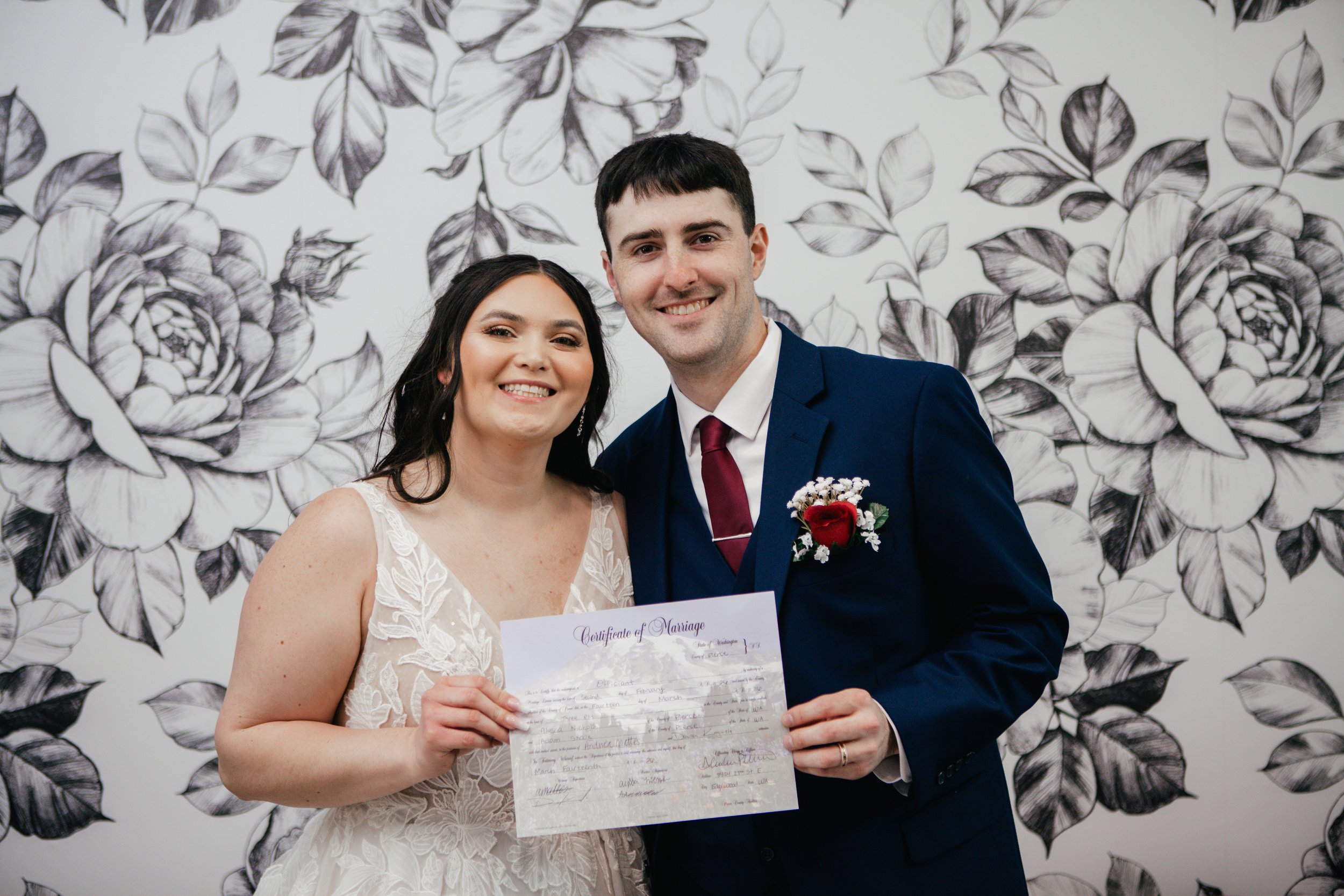 A smiling bride and groom holding a marriage certificate, standing in front of a black and white floral wall.