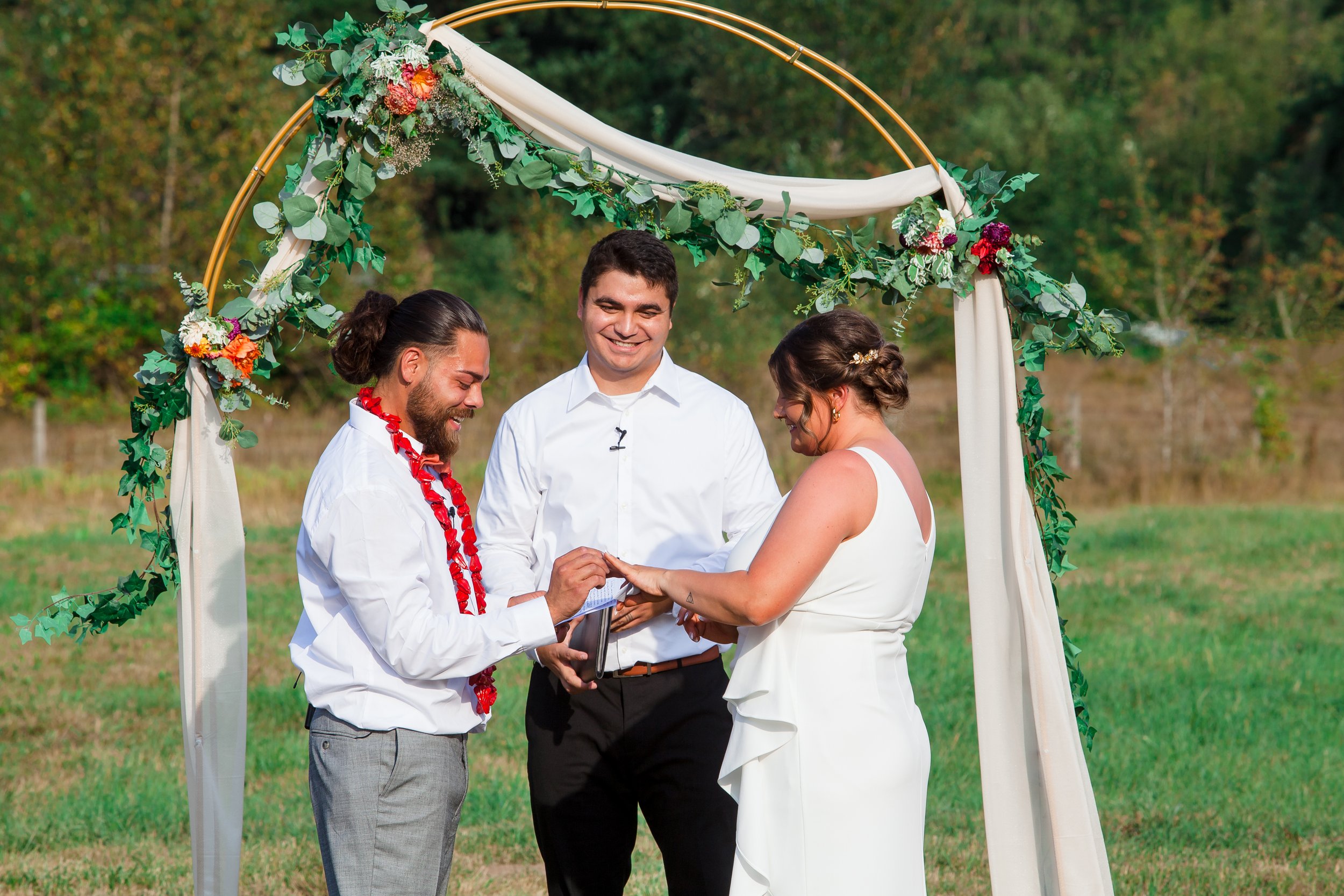 A bride and groom exchange rings during their outdoor wedding ceremony, with an officiant standing between them. The groom wears a white shirt with a red flower lei, and the bride a sleeveless white gown, beneath a circular arch decorated.