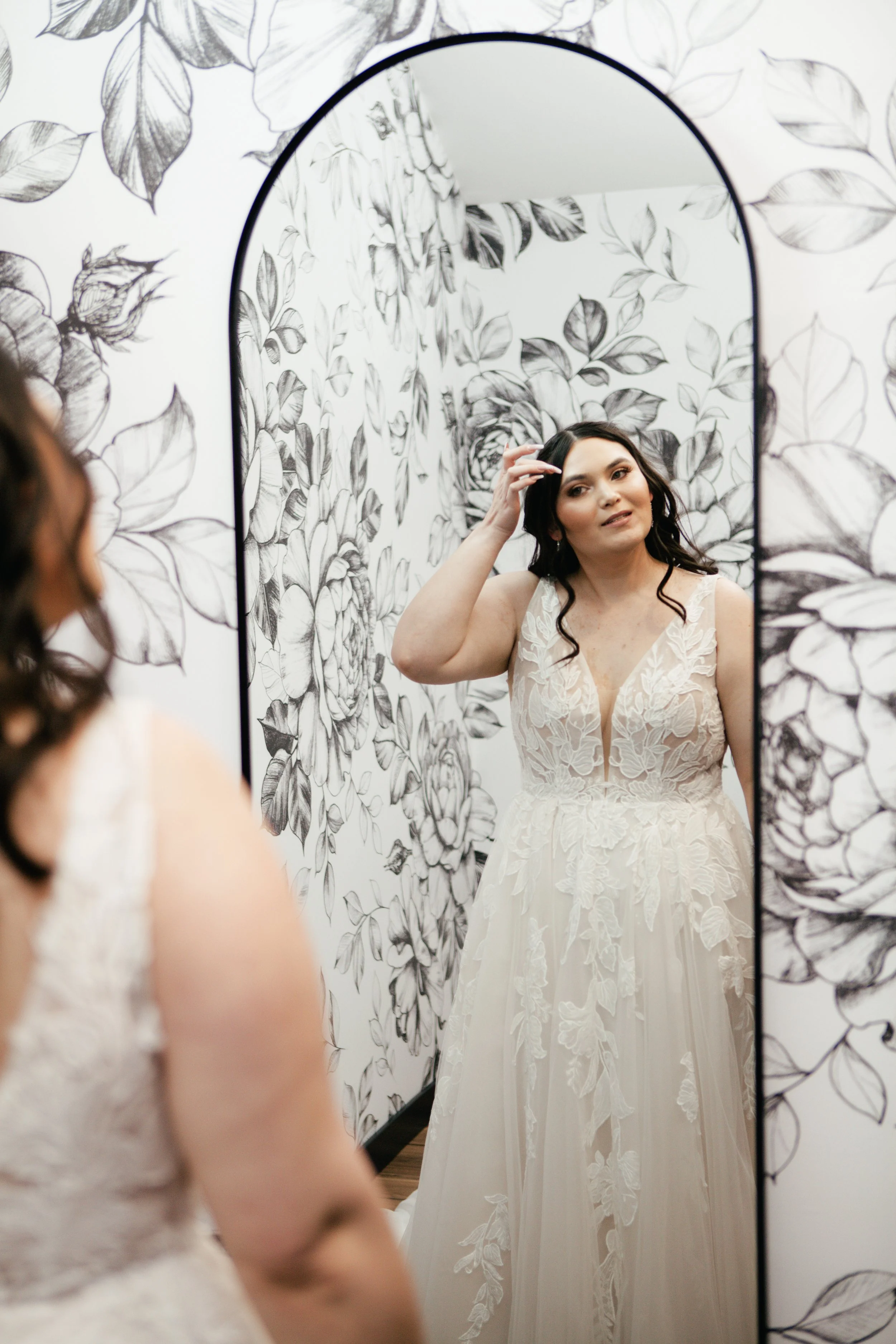 A woman in a wedding dress looking at herself in a mirror with floral wallpaper behind her.
