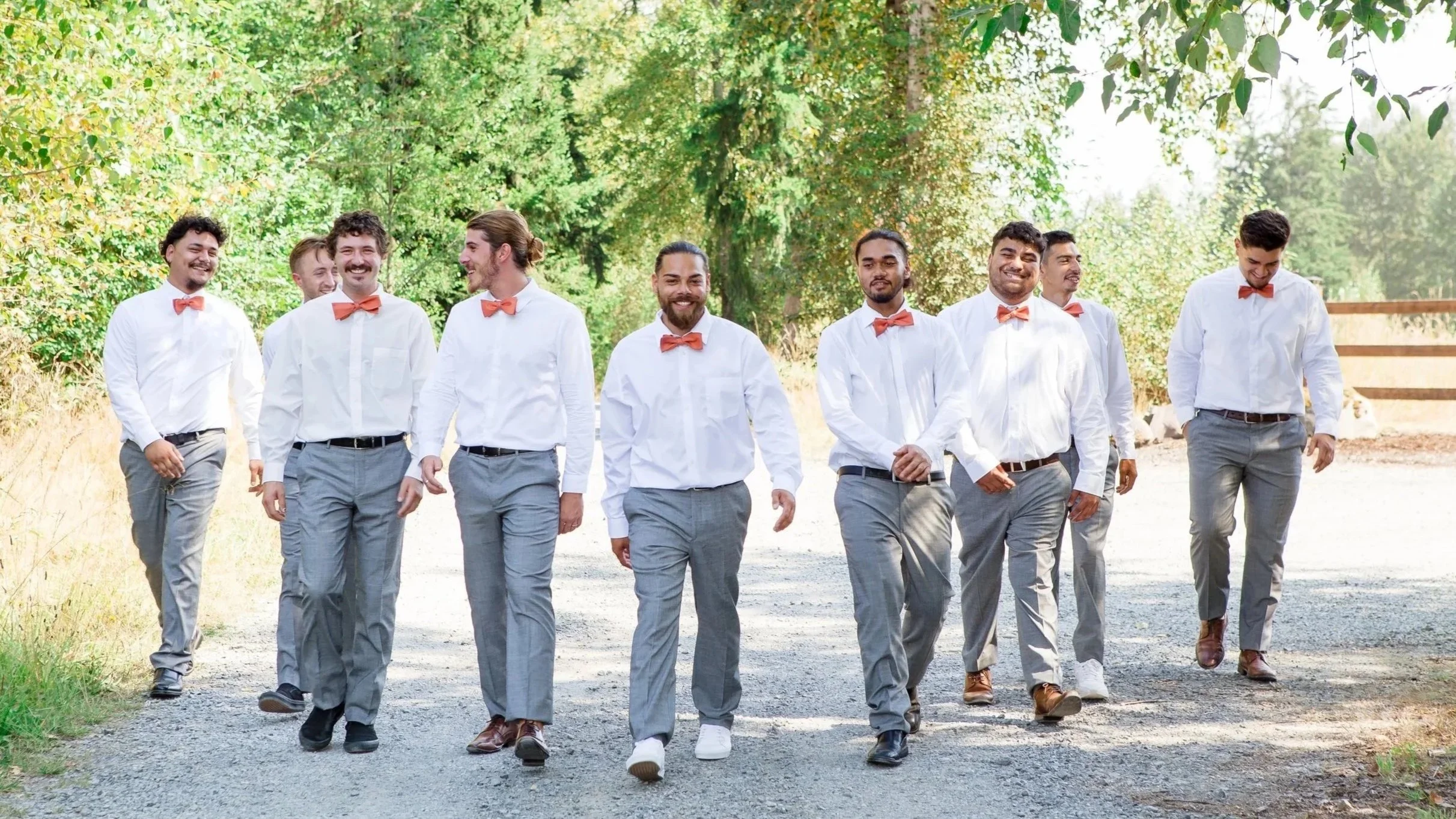 A group of men dressed in white shirts, gray pants, and orange bow ties are walking on a gravel path outdoors, surrounded by lush green trees.