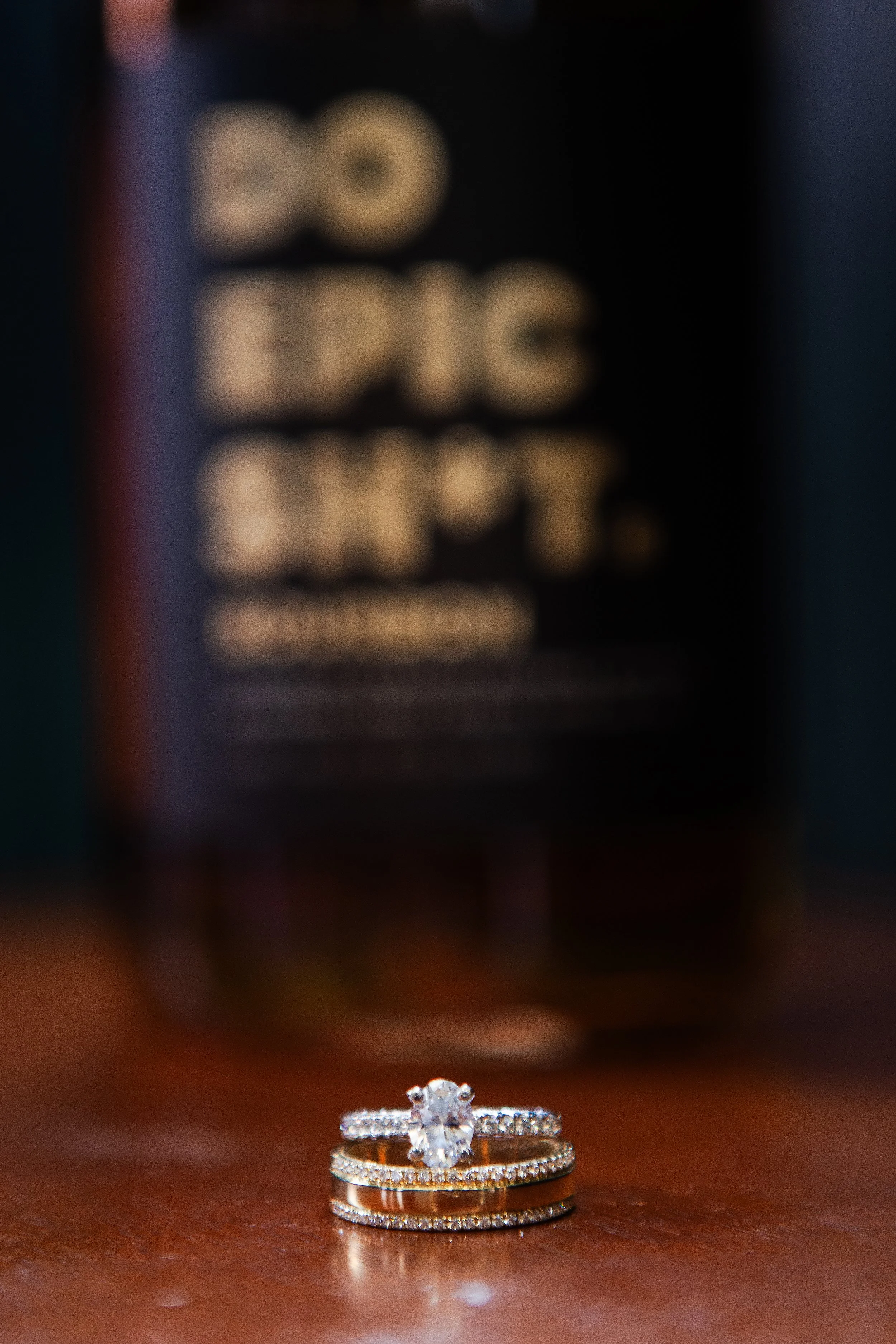 Close-up of a sparkling diamond engagement ring with a larger center diamond and two stacked wedding bands with smaller diamonds, resting on a wooden surface, with blurred rings in the background.