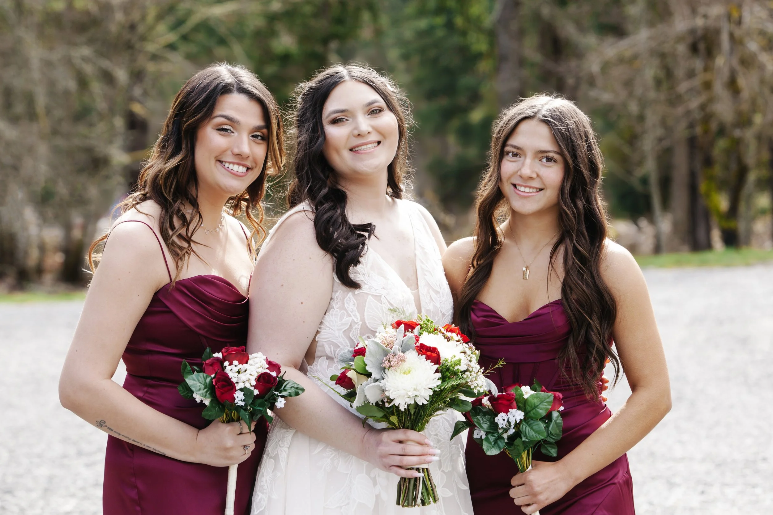 Three women dressed in formal attire standing outdoors, smiling, with one in a white dress holding a bouquet of flowers, and the other two in matching burgundy dresses, in a park-like setting with trees in the background.