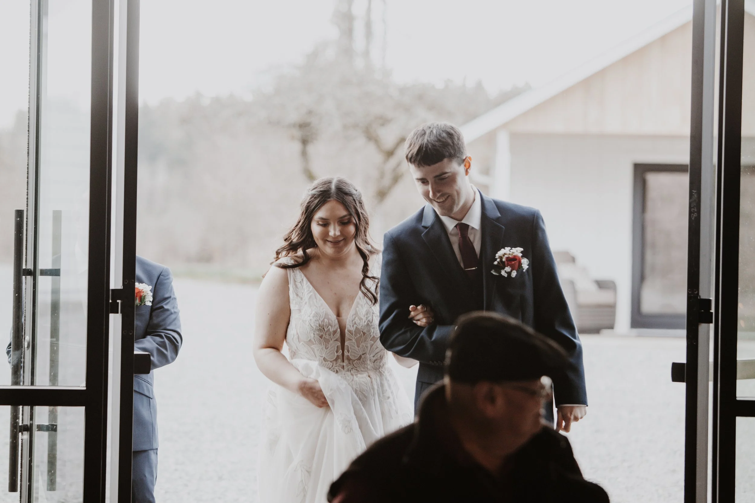 A bride and groom leaving a building, holding hands, with attendees around them. The bride is in a white lace wedding dress, and the groom is in a dark suit with a boutonniere. They are smiling and looking down, entering an outdoor area with trees an