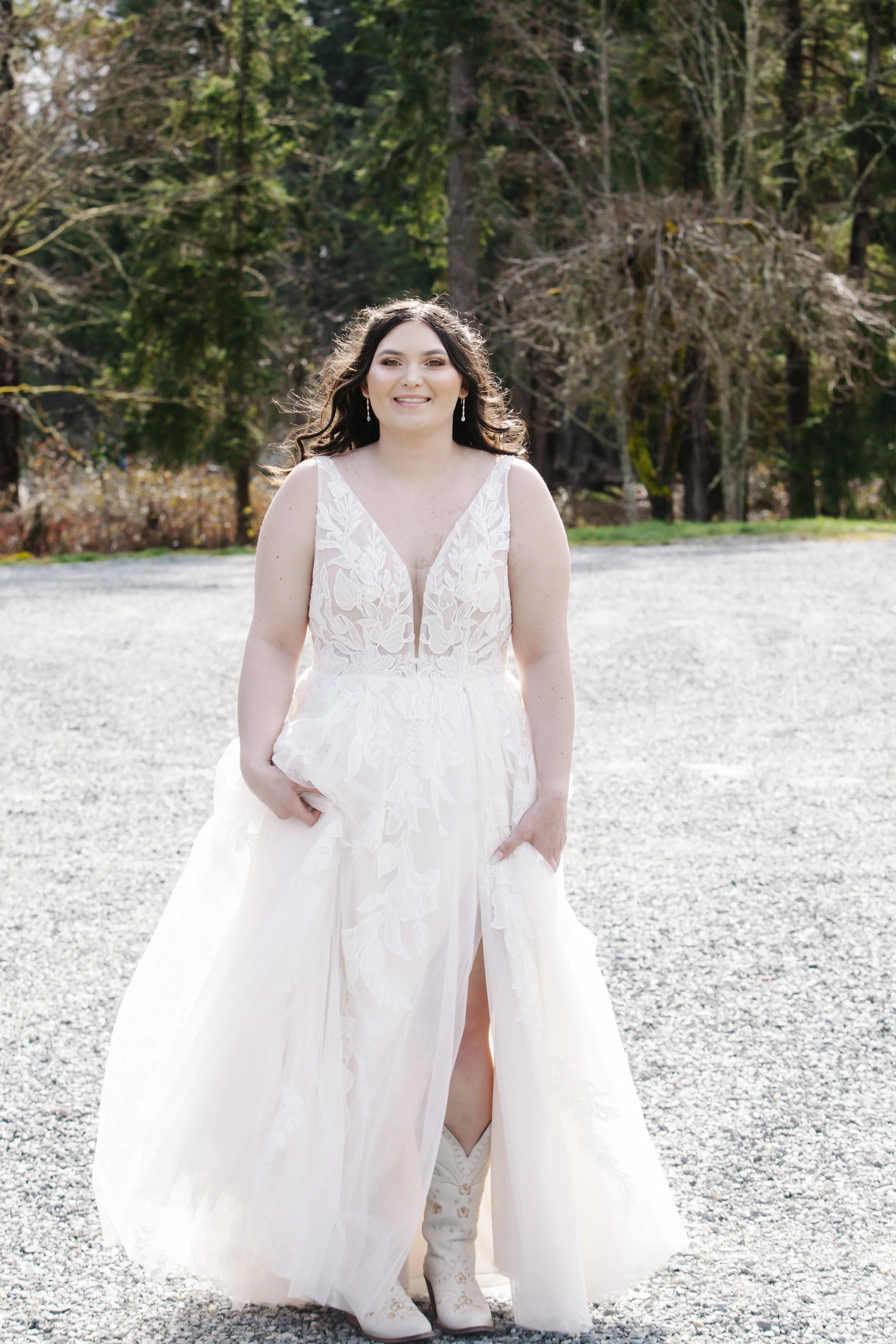 A woman in a wedding dress standing outdoors on a gravel surface with trees in the background, smiling at the camera.
