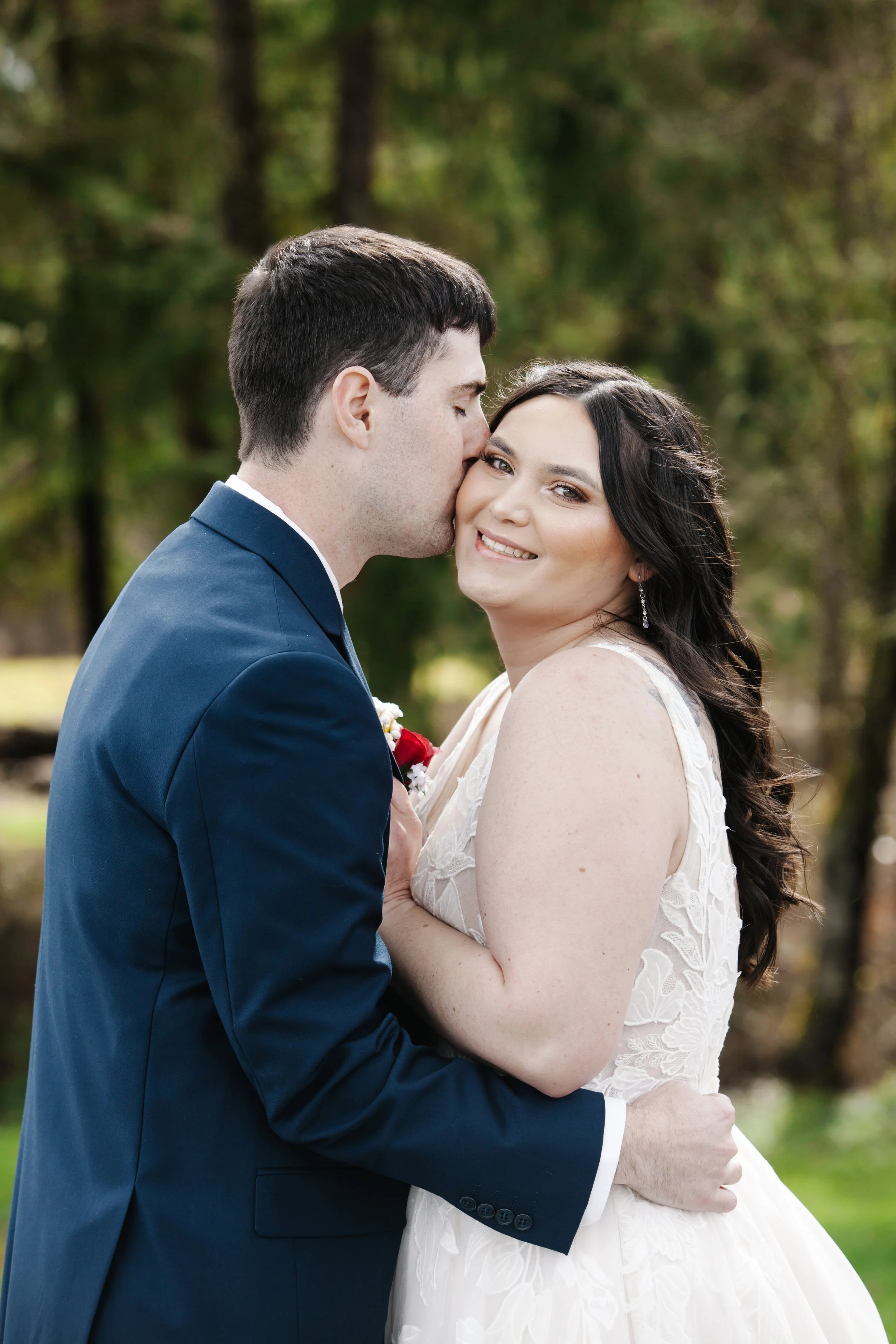 A newlywed couple outdoors, with the man in a navy suit and the woman in a white lace wedding dress, sharing an affectionate moment surrounded by greenery.