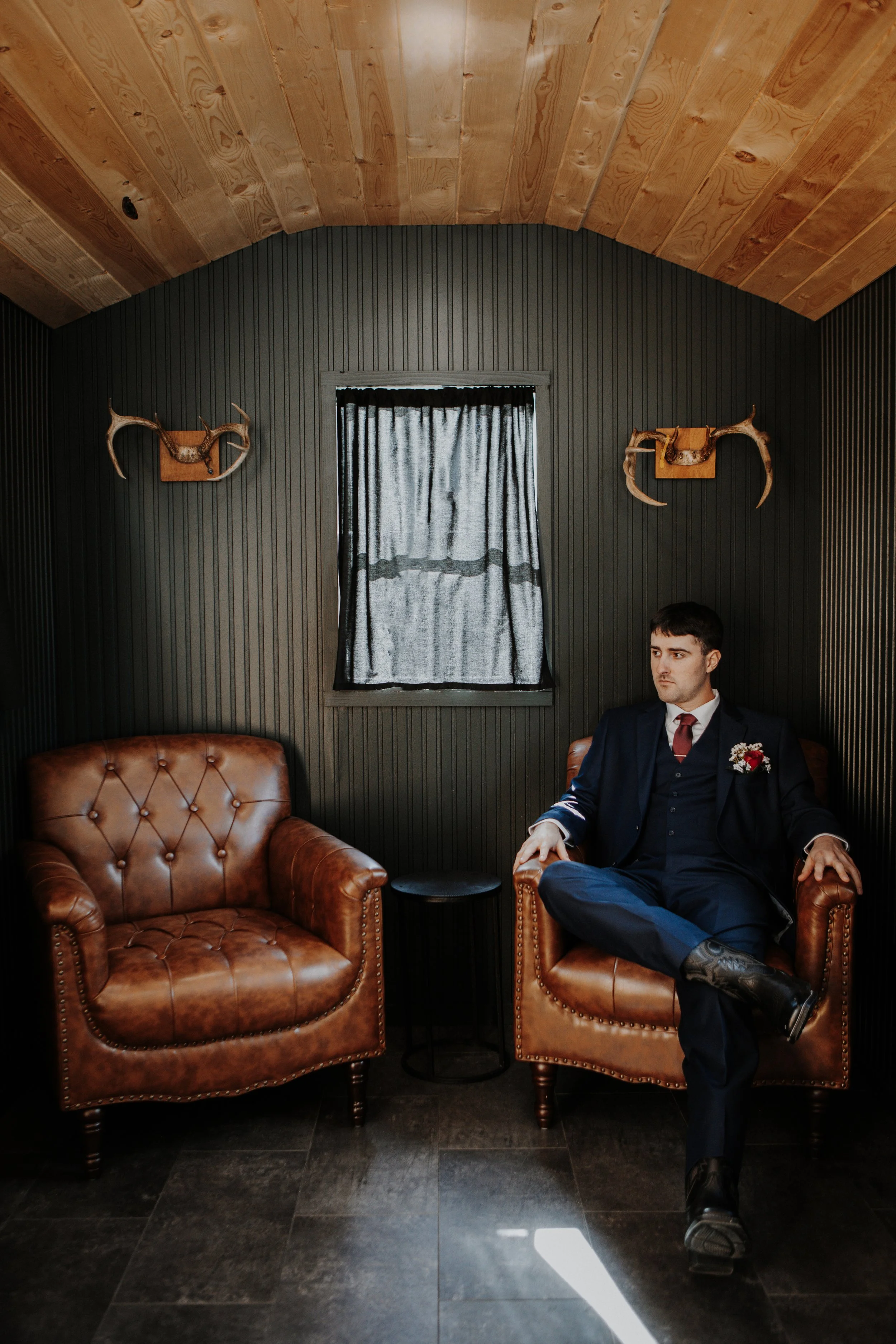 A man in a navy suit and red tie sitting on a brown leather armchair in a room with dark panel walls, a window with black curtains, and a wooden arched ceiling. There are mounted antlers on the wall and a small black side table between two chairs.