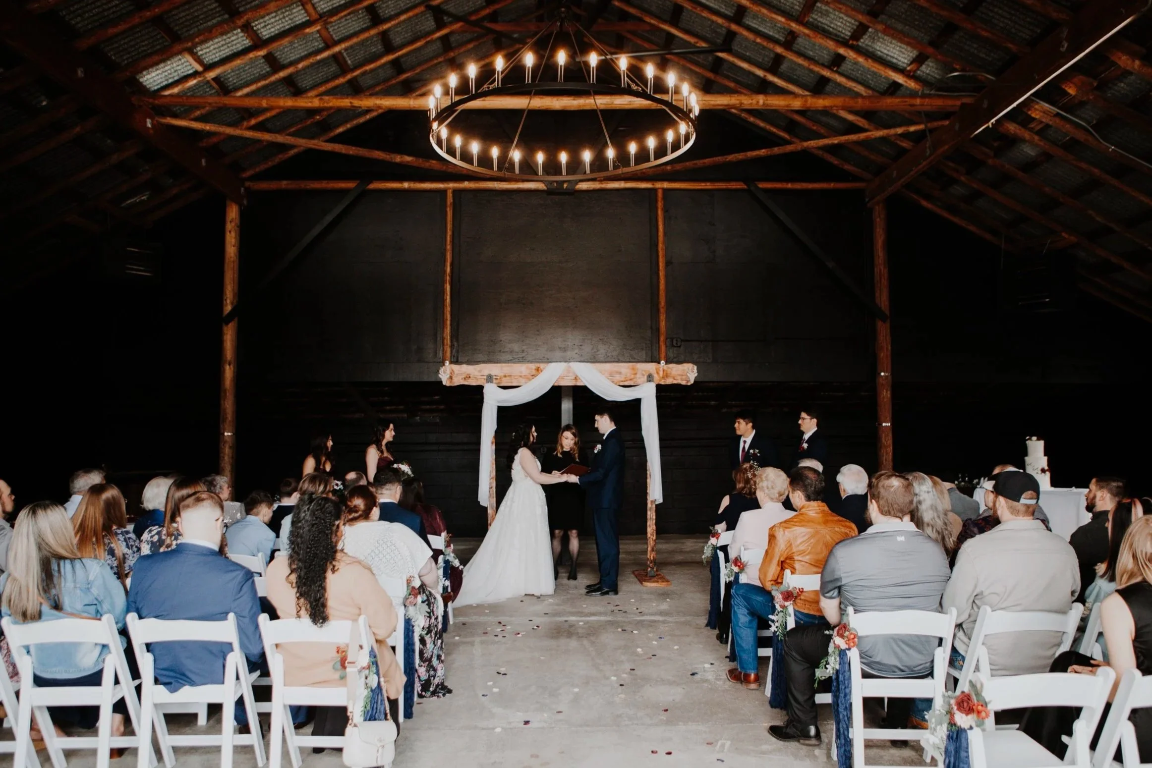 A wedding ceremony taking place inside a rustic barn with wooden beams, where a bride and groom are exchanging vows in front of an officiant, with guests seated on white chairs watching.