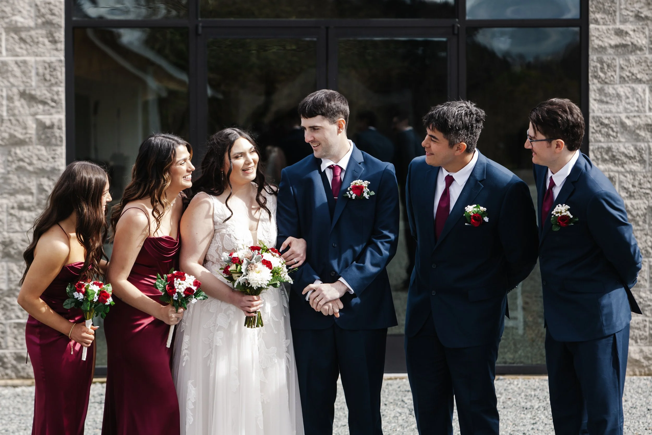 A bride and groom at a wedding ceremony, exchanging vows, surrounded by bridesmaids and groomsmen outdoors with a building in the background.