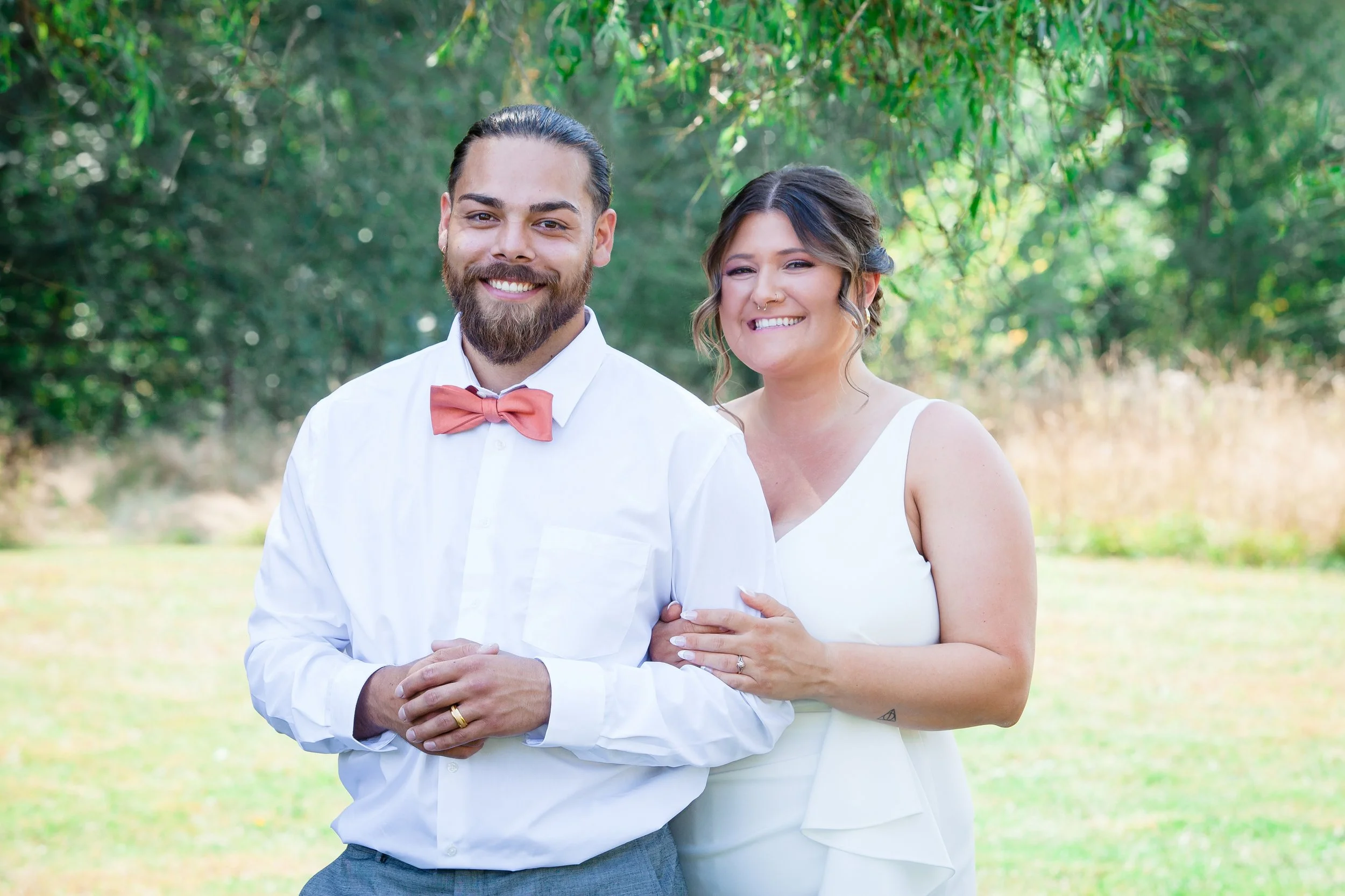 A smiling couple standing outdoors in a green, wooded area. The man has a beard, long dark hair tied back, and is wearing a white shirt with a pink bow tie. The woman has curly hair with highlights, a nose ring, and is dressed in a white sleeveless dress. They are standing close together, smiling happily.