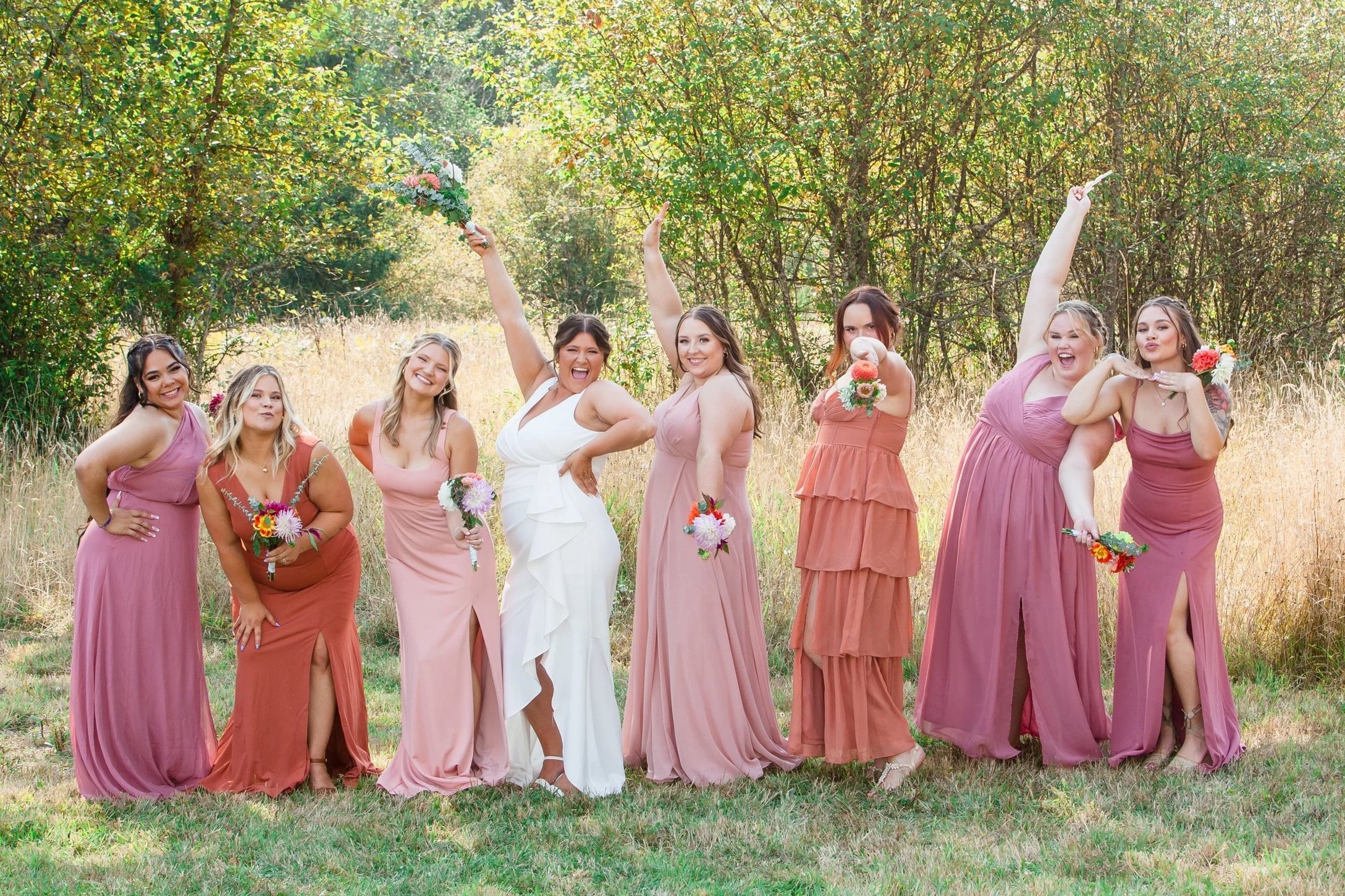 Group of women in pastel-colored dresses celebrating outdoors, some holding bouquets, in a grassy area with trees in the background.