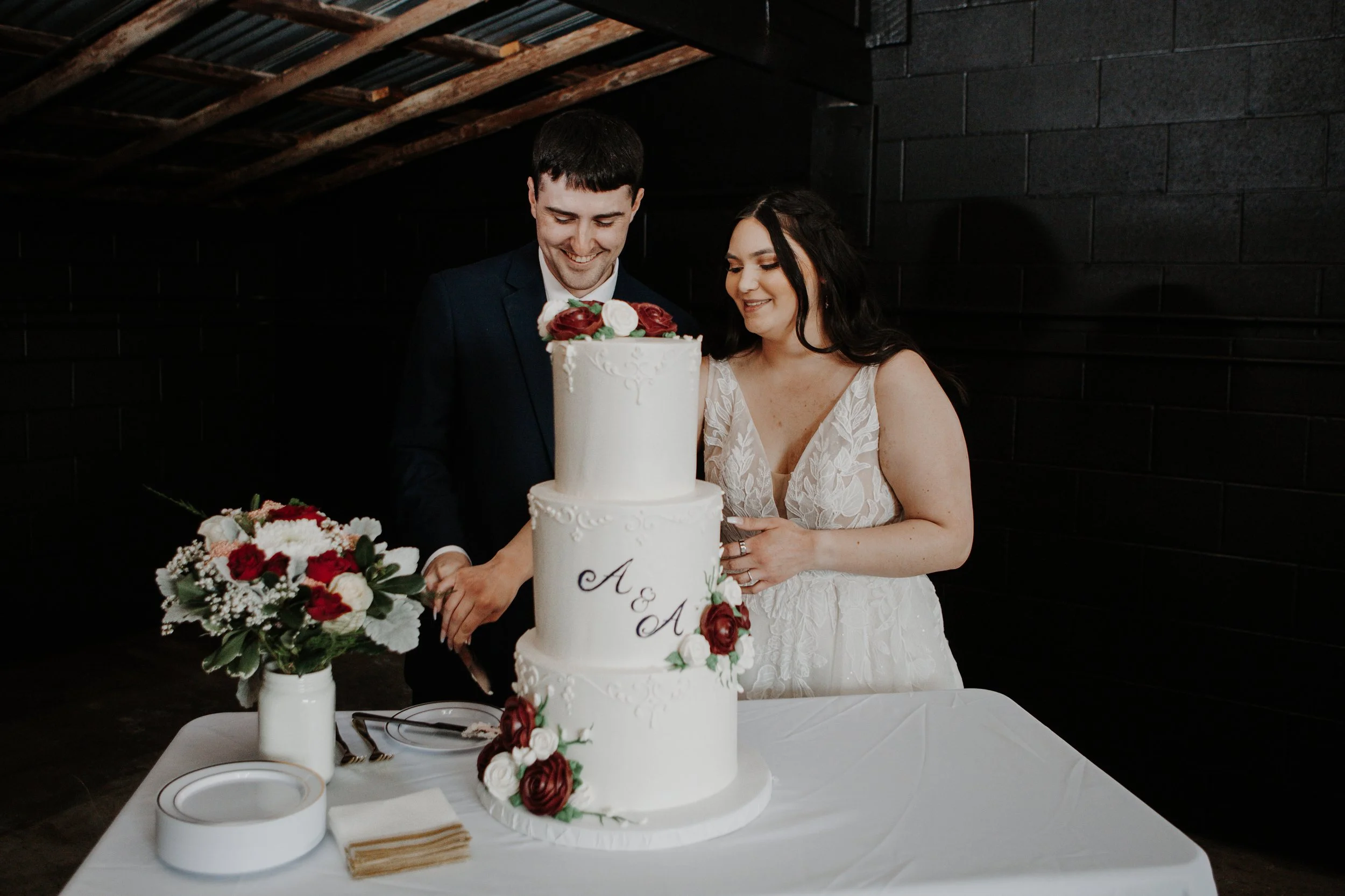 A bride and groom cutting a white wedding cake with red and white flowers on top and around the base, with a bouquet of red and white flowers on the table beside them, in a dark room with a black brick wall.