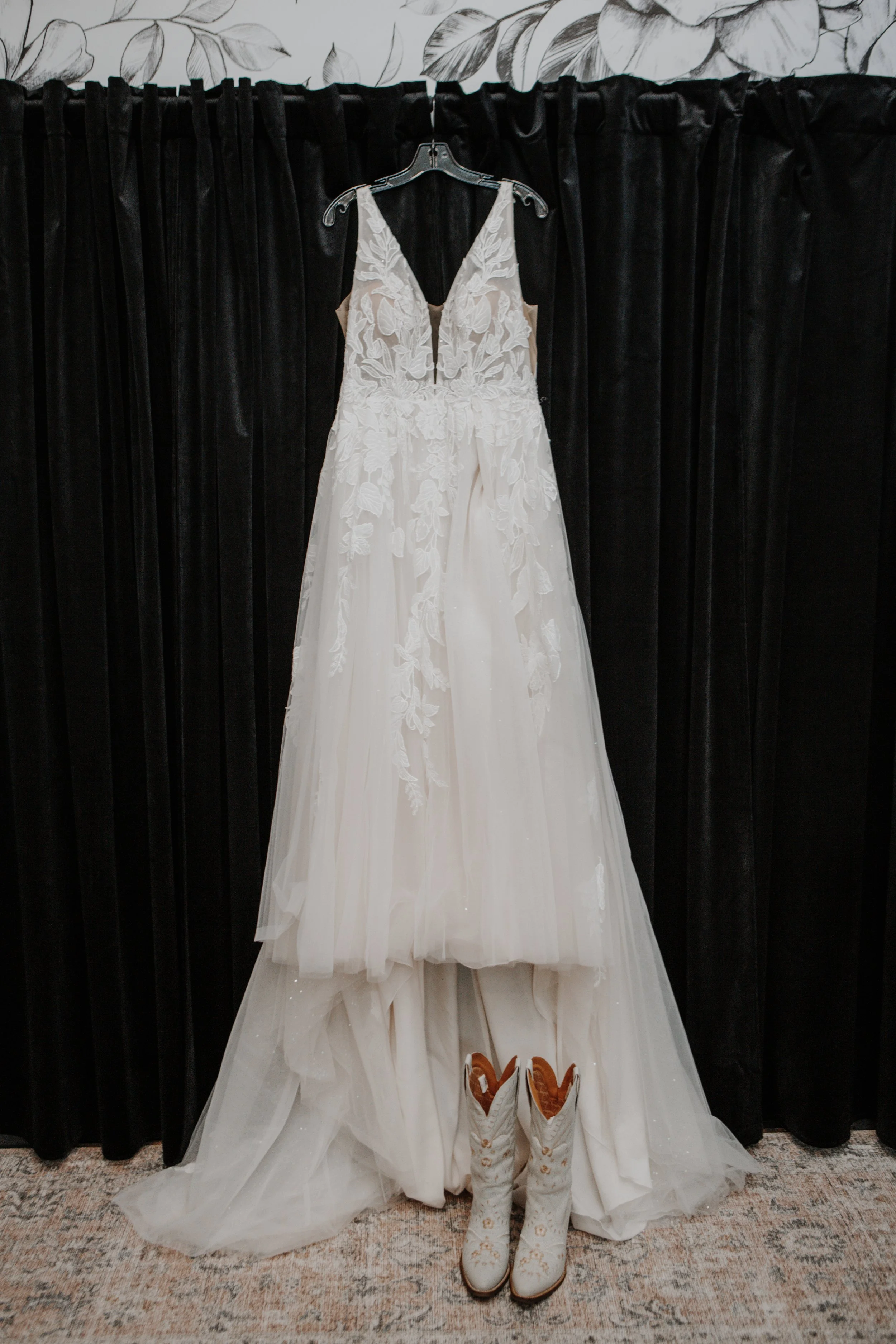 A wedding dress hanging on a hanger in front of a black curtain, with cowboy boots placed at the bottom of the dress on a patterned carpet.