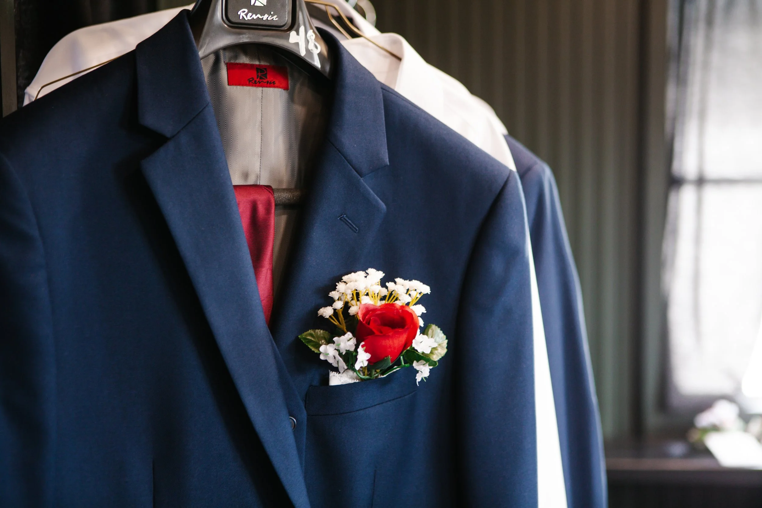 A navy blue suit jacket with a red boutonniere of flowers pinned to the pocket, hanging on a black hanger labeled 'Renoir'.