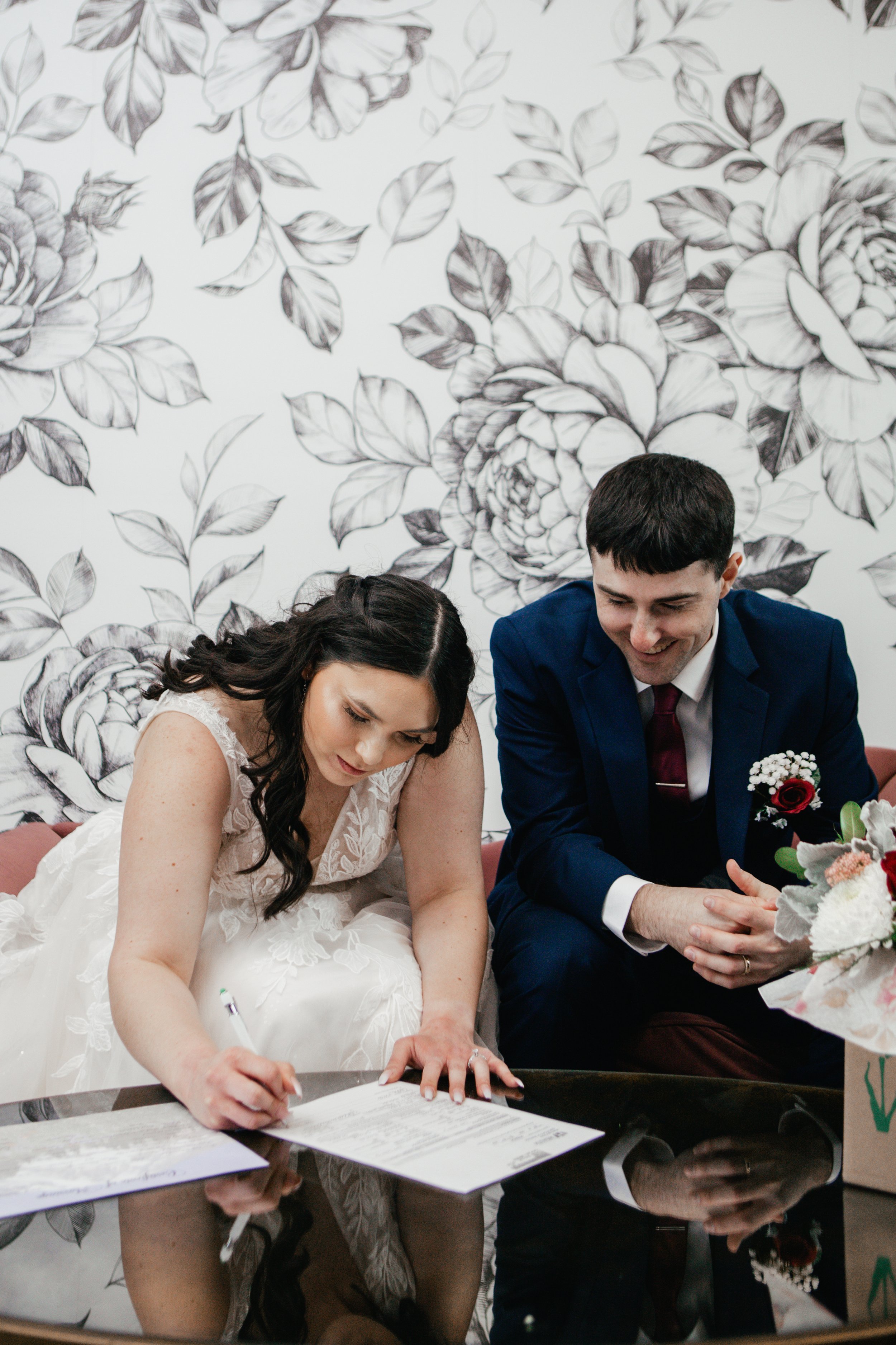 A bride with long dark hair and wearing a white lace wedding dress signing a document, and a groom with short dark hair wearing a dark suit, white shirt, and red tie, sitting beside her with clasped hands and a red rose boutonniere, both smiling duri