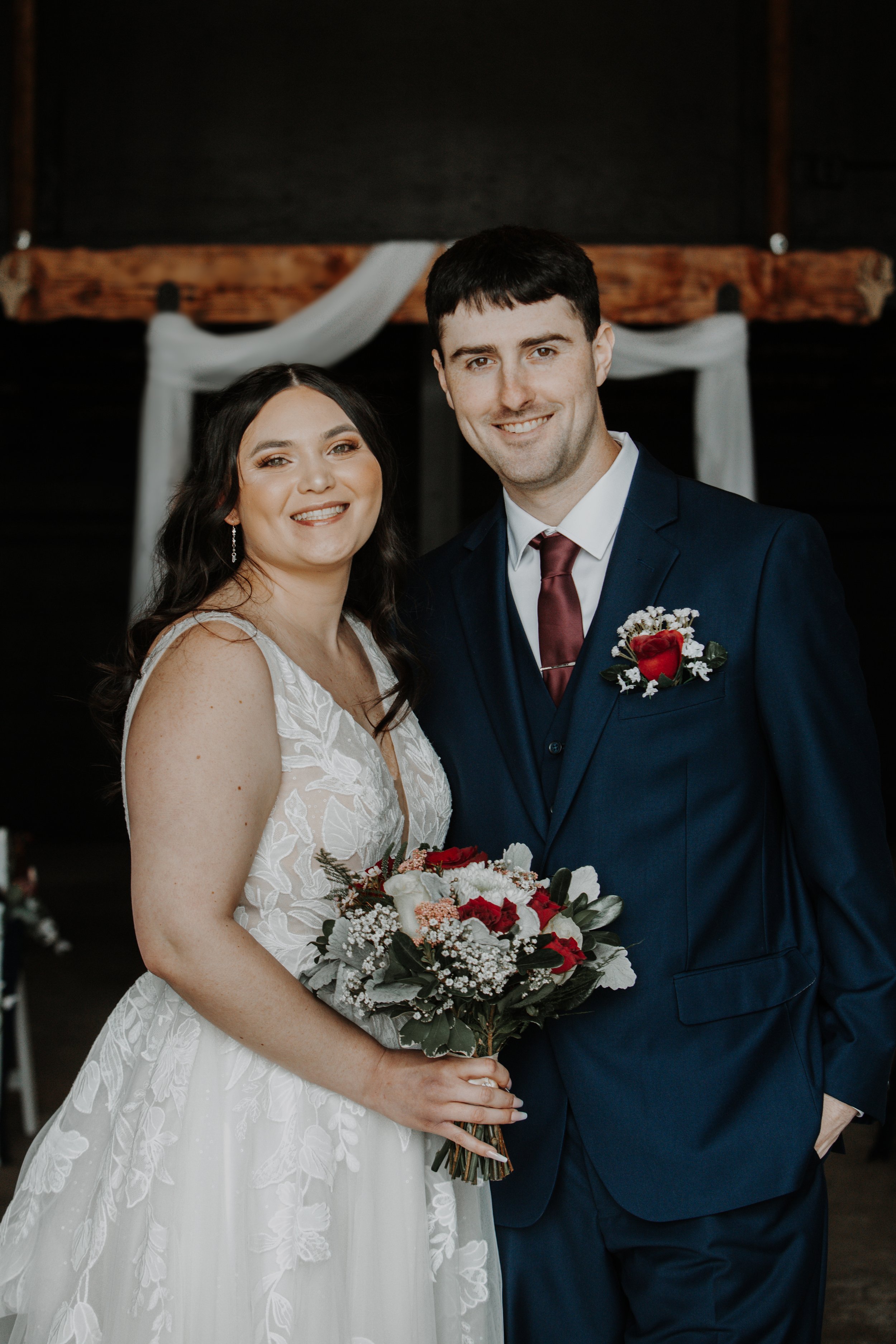 A bride and groom smiling at each other during their wedding ceremony, with the bride holding a bouquet of red, white, and pink flowers and the groom dressed in a navy suit with a red tie and a red boutonniere, standing under a wooden arch decorated 
