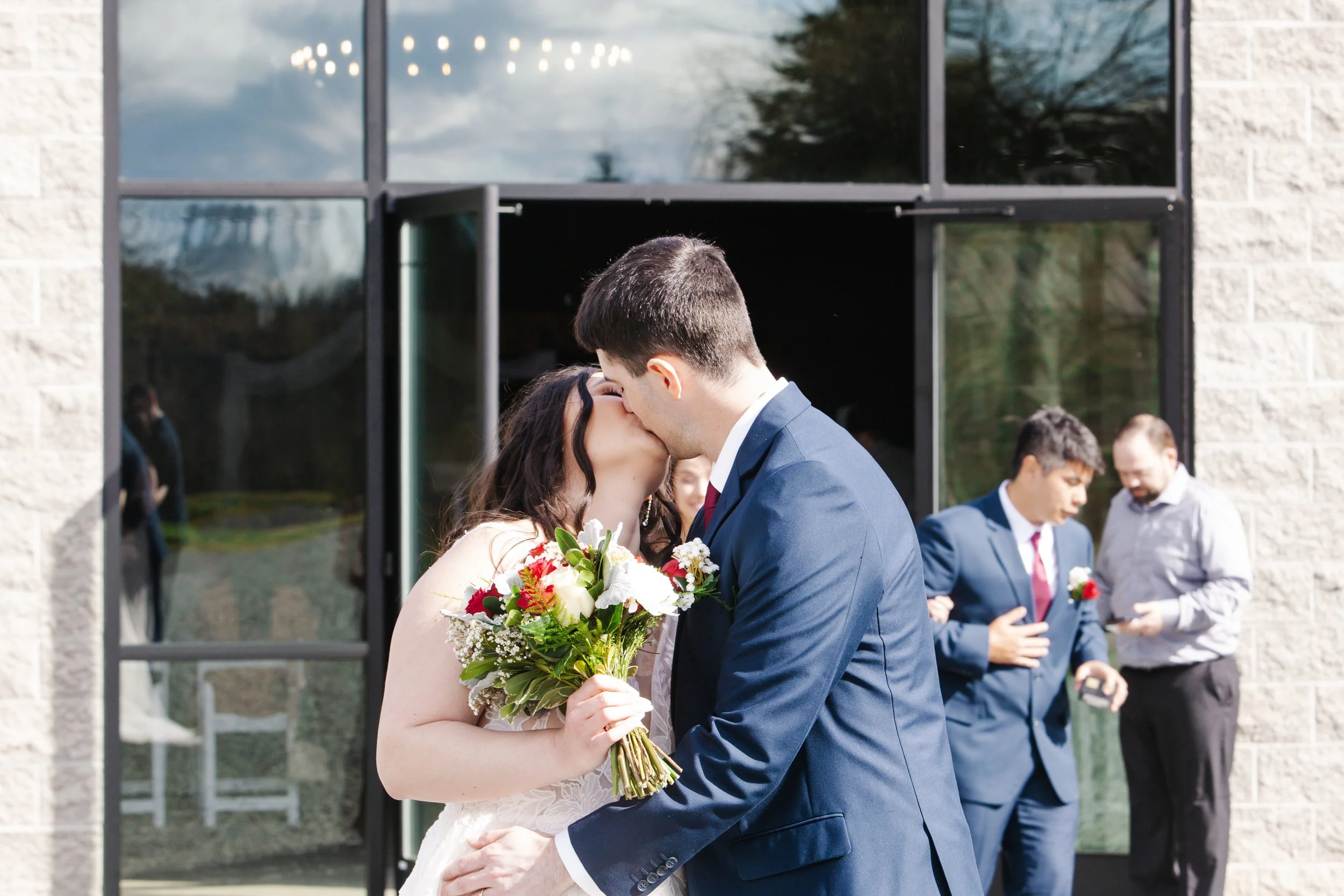 A bride and groom share a kiss outside a building during their wedding ceremony, with the bride holding a bouquet of flowers and the groom dressed in a navy suit.