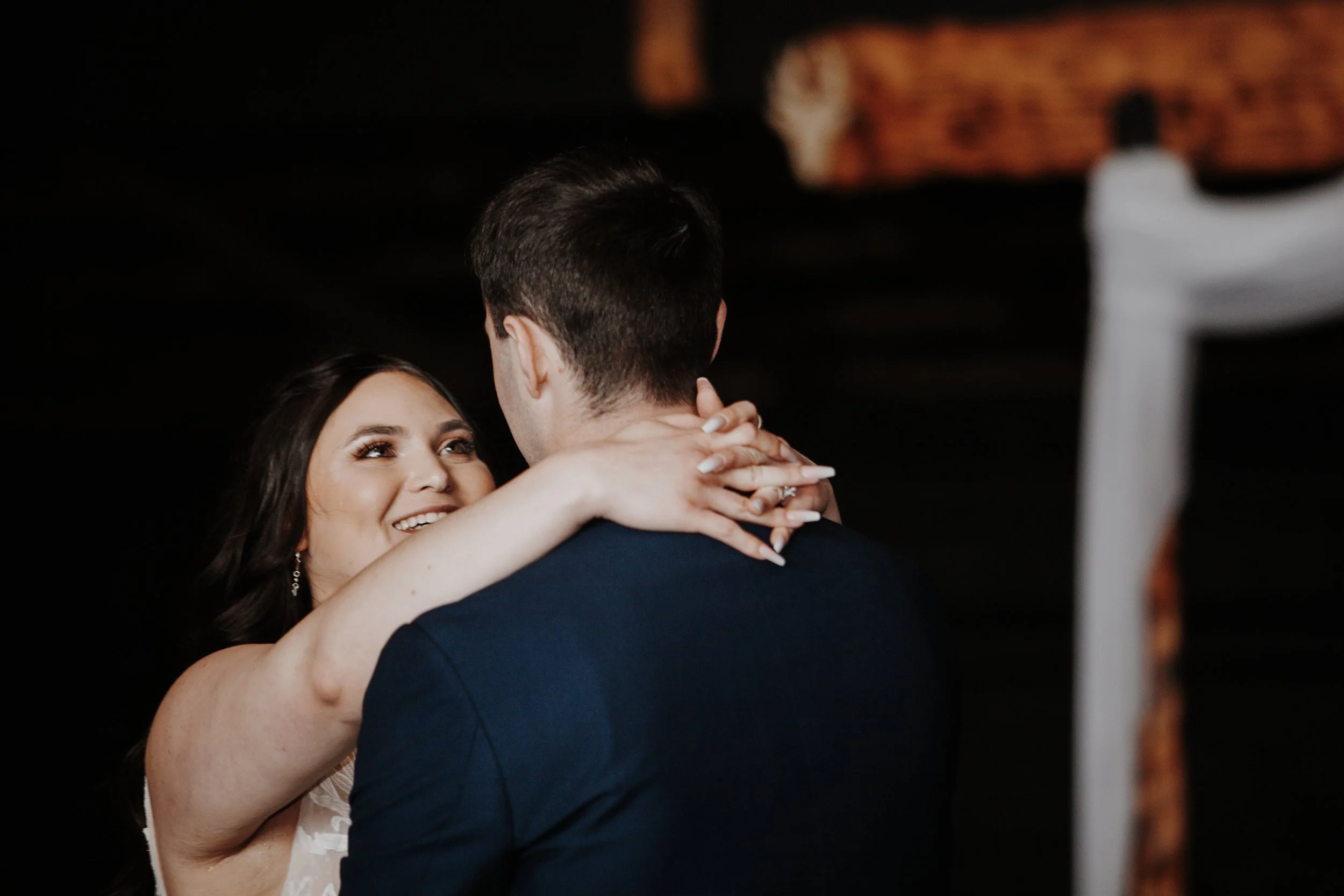 A woman in a white dress and a man in a dark suit share a dance, looking into each other's eyes in a dimly lit setting.
