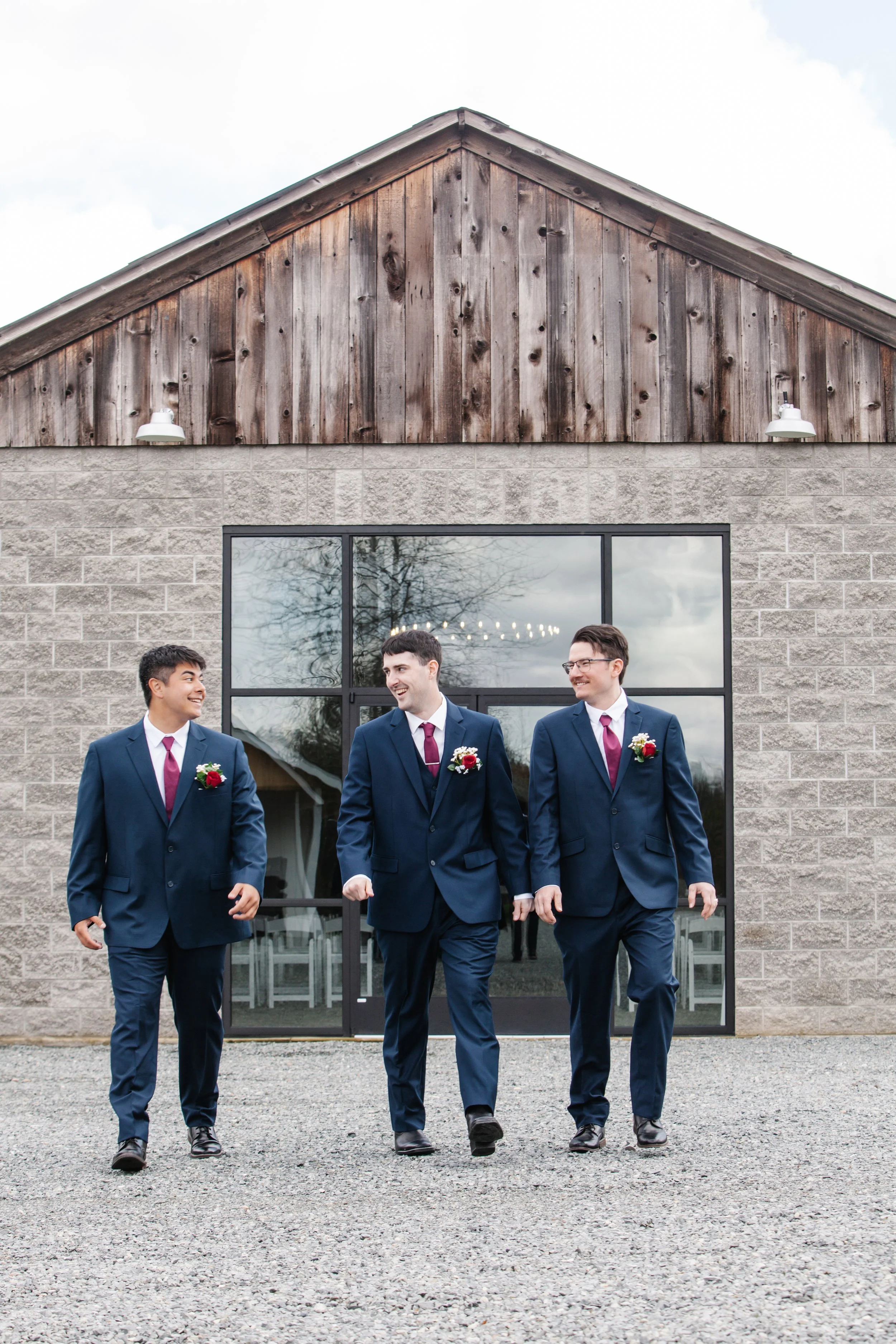 Three young men in blue suits with boutonnieres walking together outside a building with a rustic wood and brick facade.