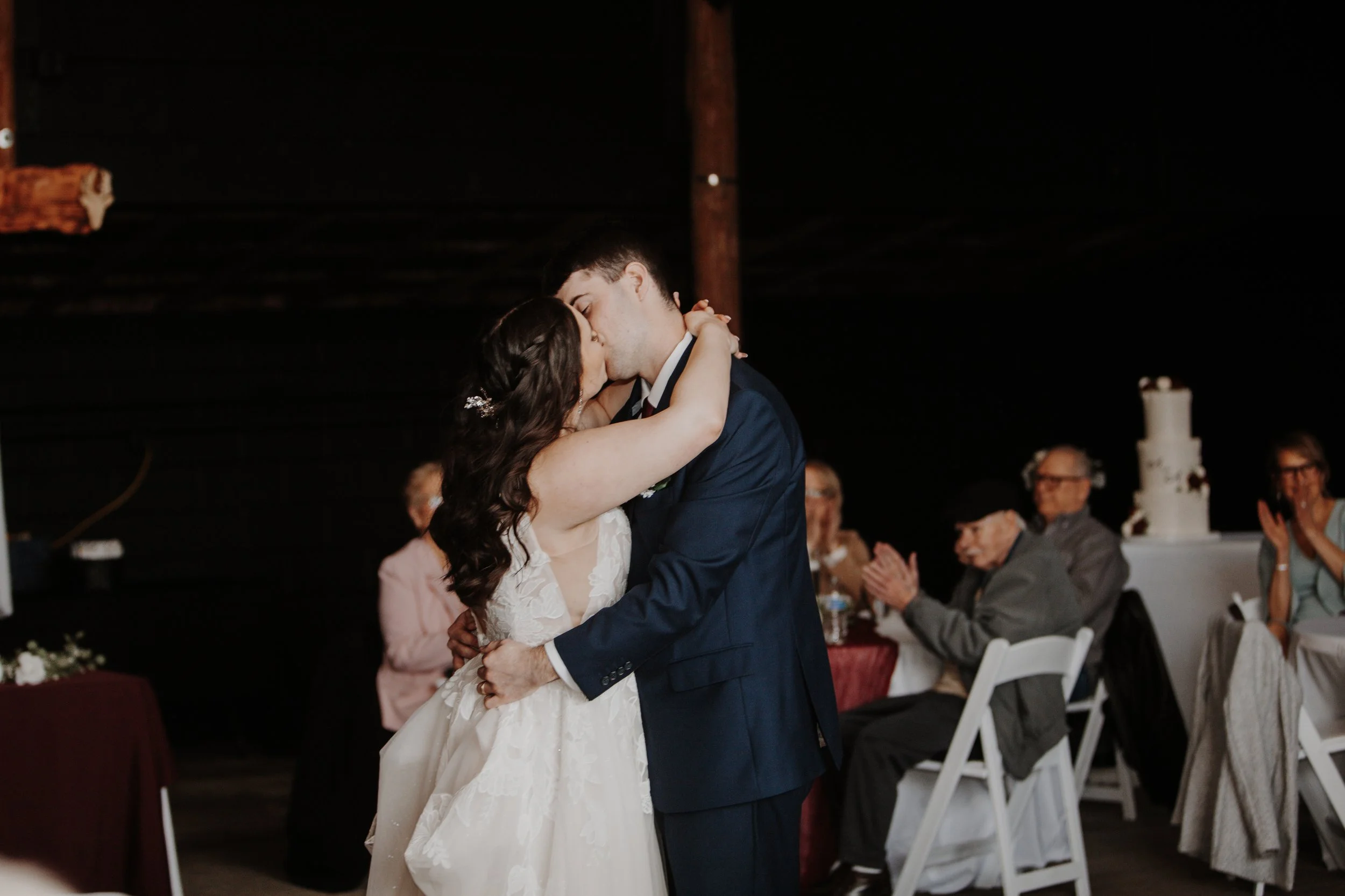 A bride and groom sharing a kiss during their wedding dance, surrounded by seated guests clapping, inside a dimly lit venue.