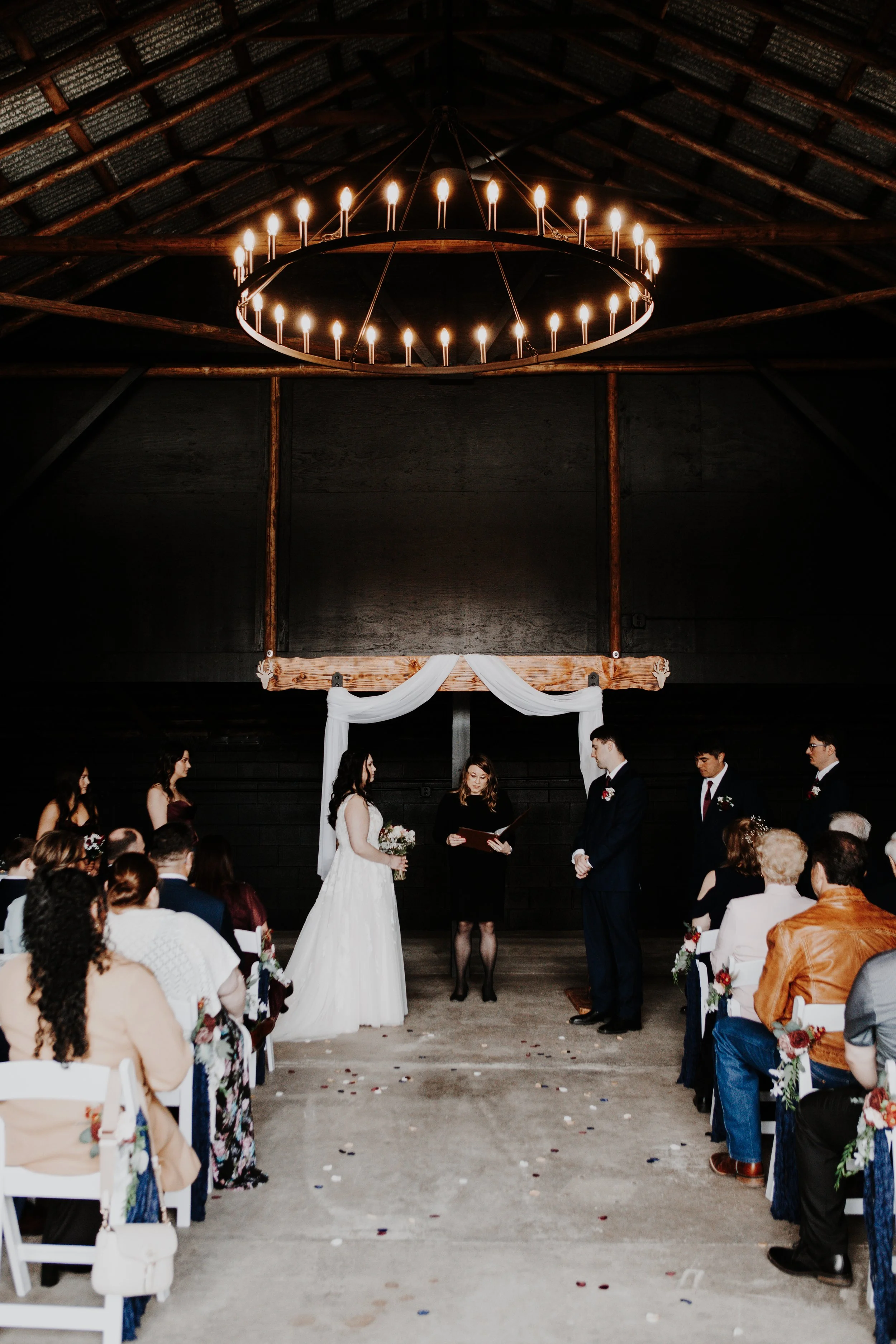 A wedding ceremony taking place indoors with guests seated on both sides of an aisle decorated with confetti and flower accents. The couple is standing in front of an officiant, with the bride in a white gown holding a bouquet and the groom in a dark