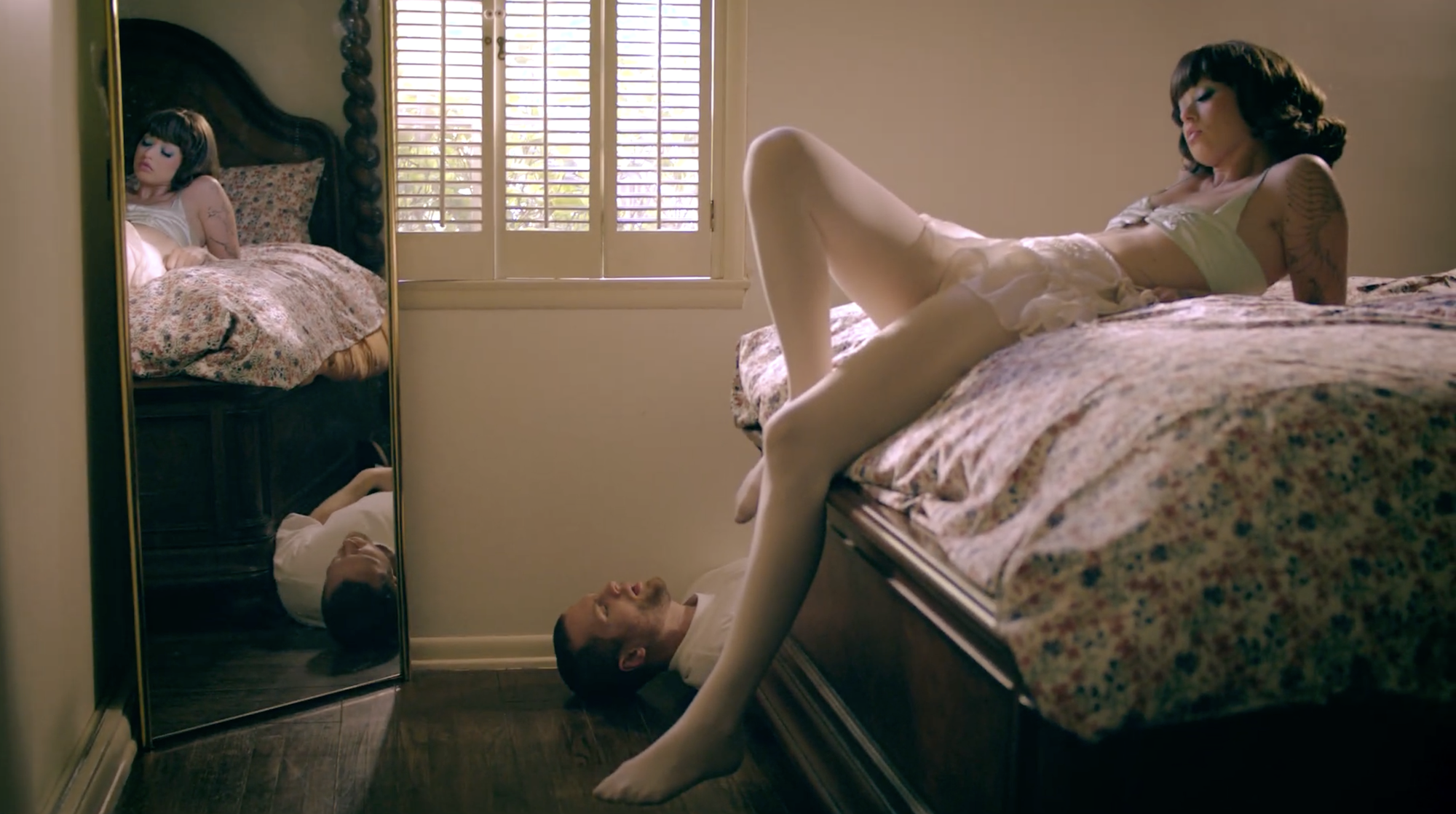 A woman lying on a bed with her head turned to the side and eyes closed, and a man lying underneath the bed with his head near her legs, looking up with eyes closed. The scene is reflected in a mirror on the wall beside the bed, with a window with shutters in the background.
