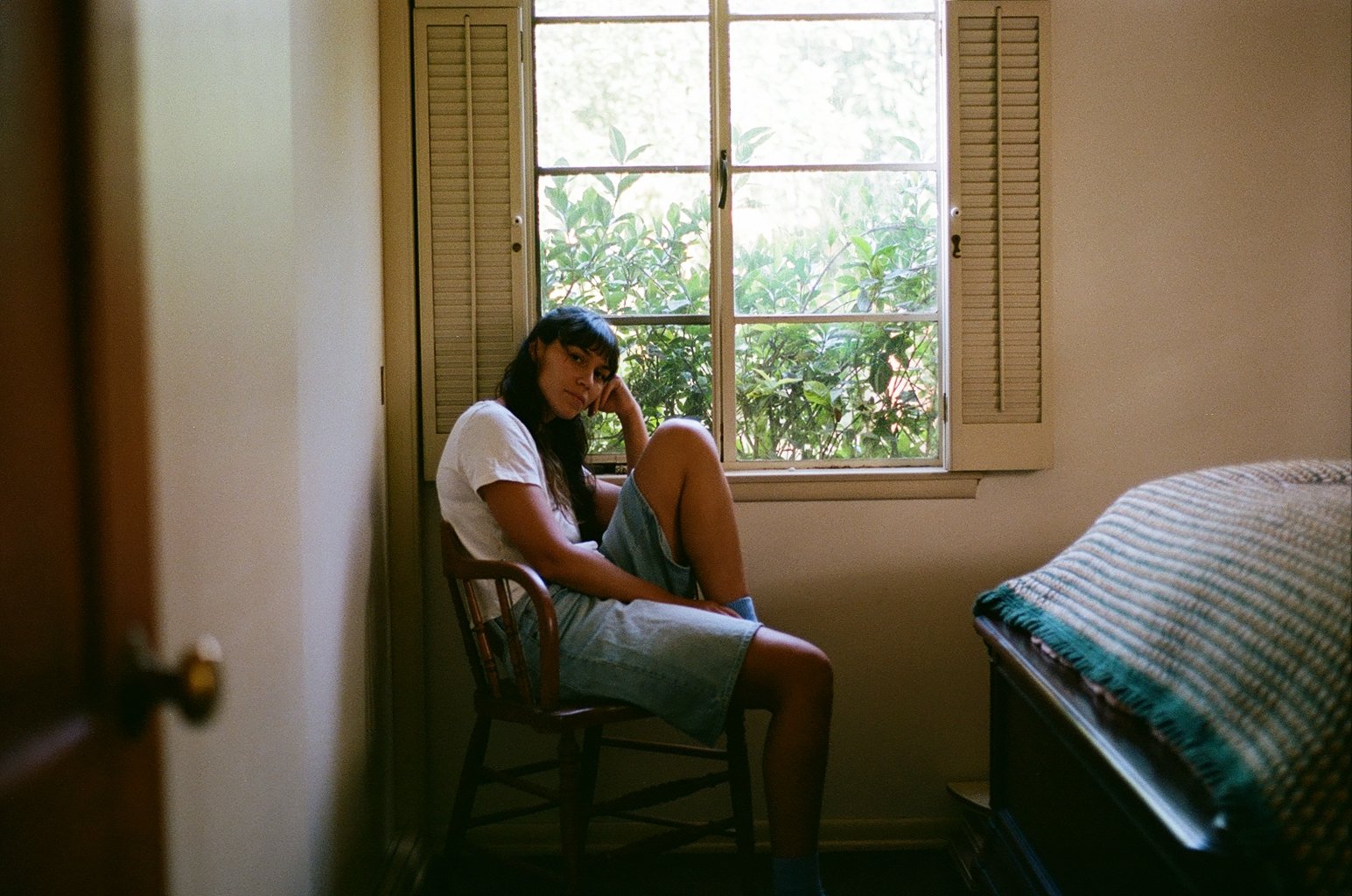 Young woman with long dark hair sitting on a wooden chair in a room with a window and green foliage outside. She is wearing a white t-shirt and shorts, resting her head on her hand, with a contemplative expression.