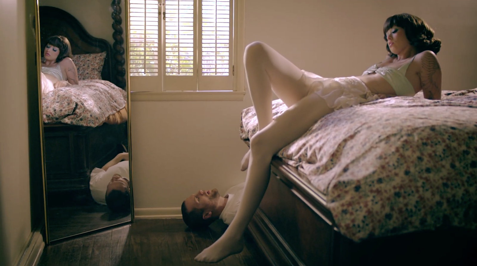 A woman with dark hair and tattoos lying on a bed, and a man lying on the floor with his head under the bed, creating an optical illusion with the woman's legs.