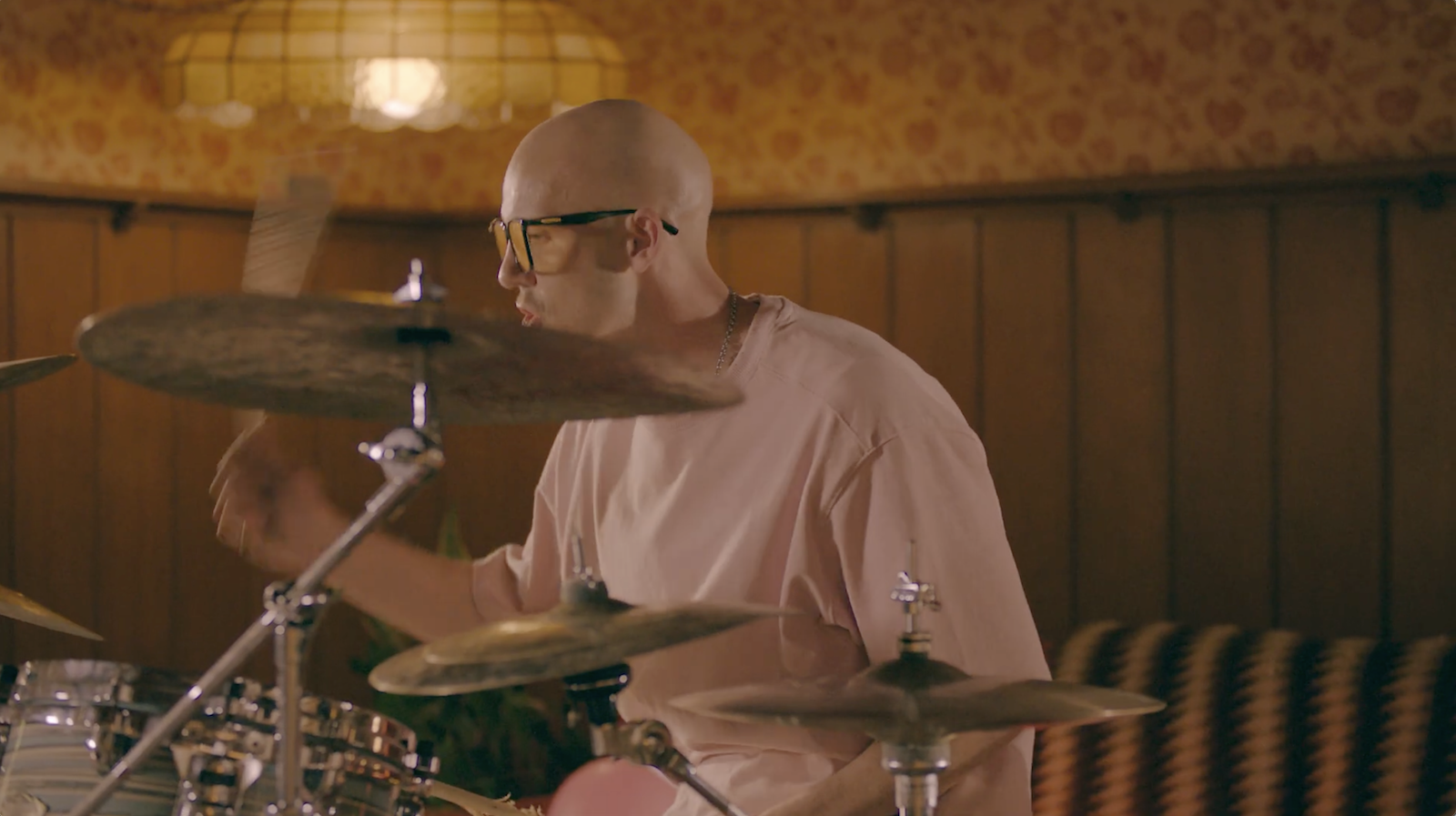 A man with a shaved head and glasses playing drums in a room with wood-paneled walls and vintage wallpaper.