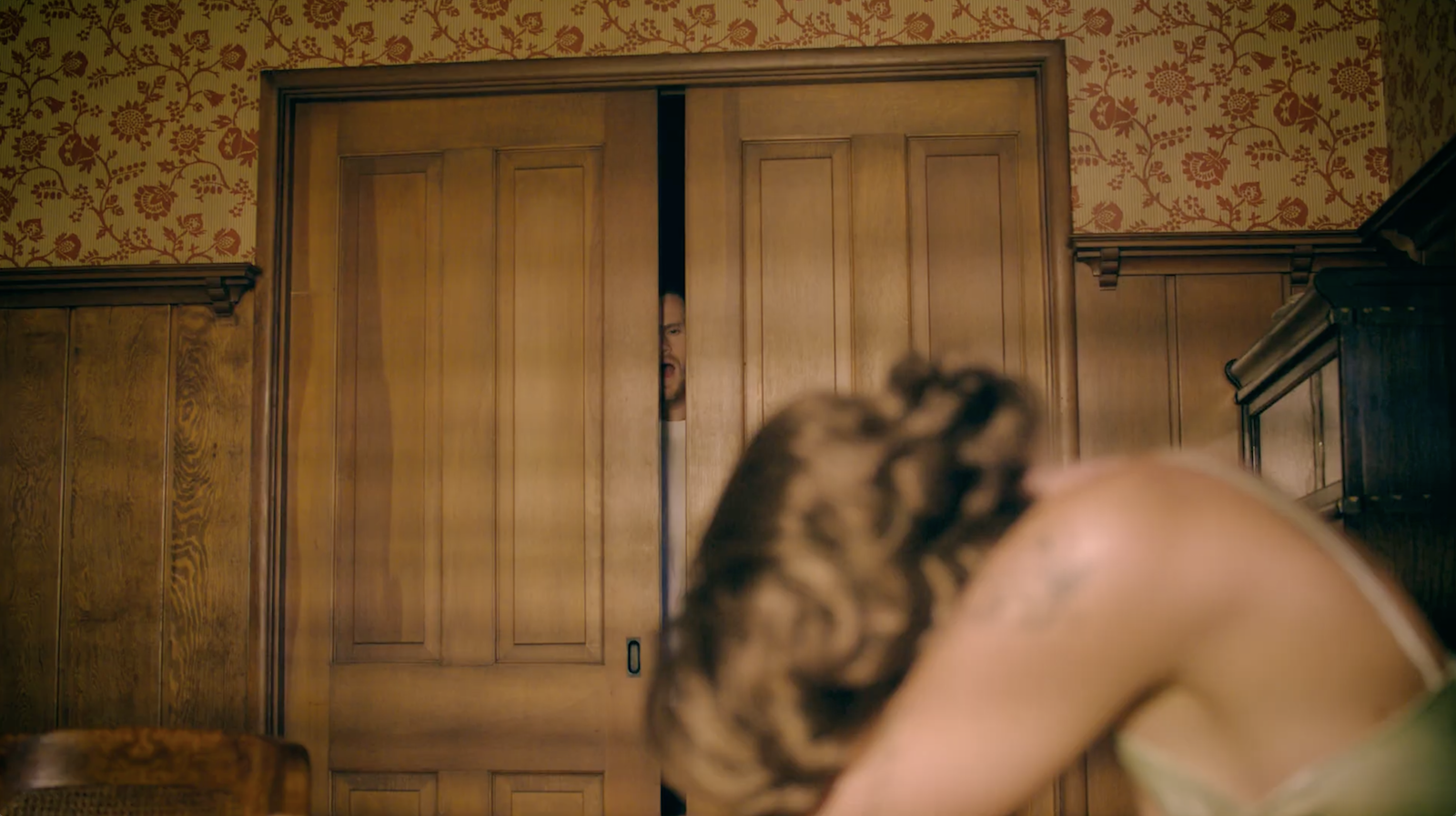 Person with curly hair in the foreground, bending forward, looking toward a person peeking through a partially open wooden door in the background.