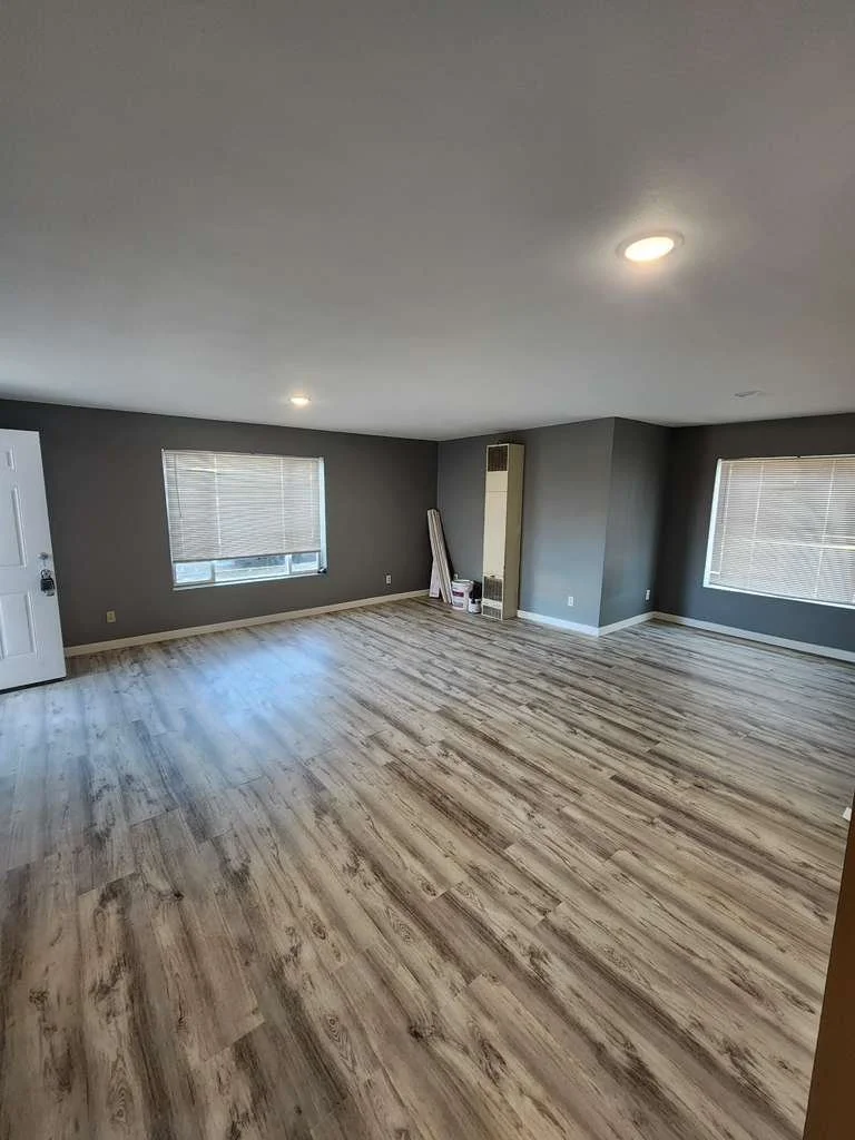 Empty living room with gray walls, large windows with blinds, light wood flooring, and a ceiling light.