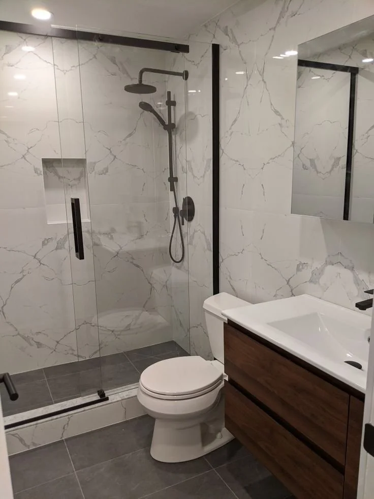 Modern bathroom with a walk-in shower enclosed by glass, featuring black fixtures and marble walls, a toilet, and a wooden vanity with a white sink.