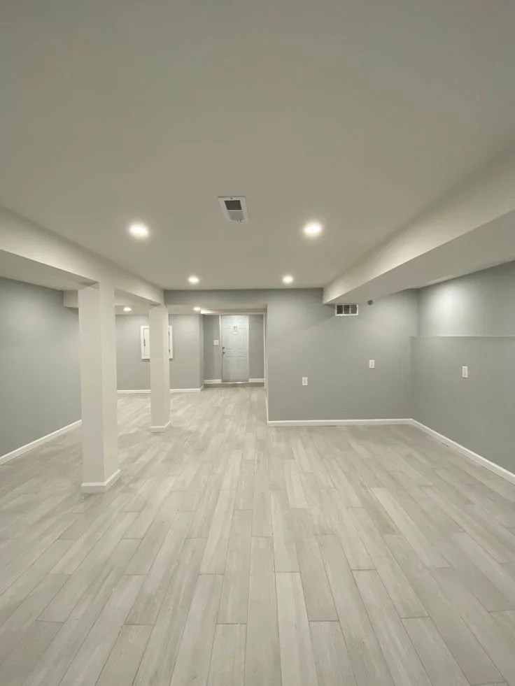 Empty basement room with light gray walls, white trim, and light wood flooring, illuminated by ceiling lights.
