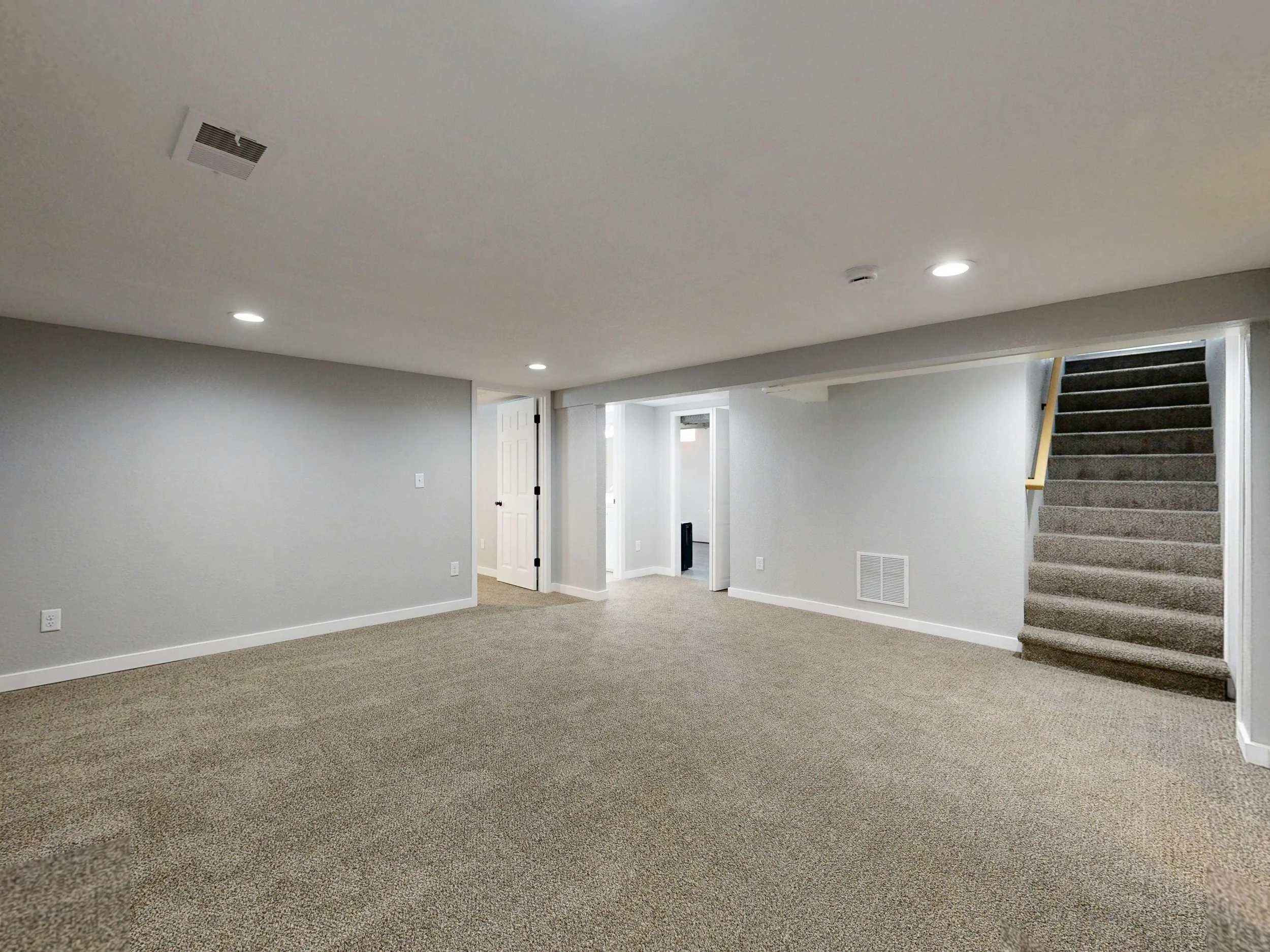 A spacious, empty basement with beige carpet, light gray walls, and a white ceiling with recessed lighting. There is a staircase with dark gray carpet leading upstairs on the right and a white door on the left.