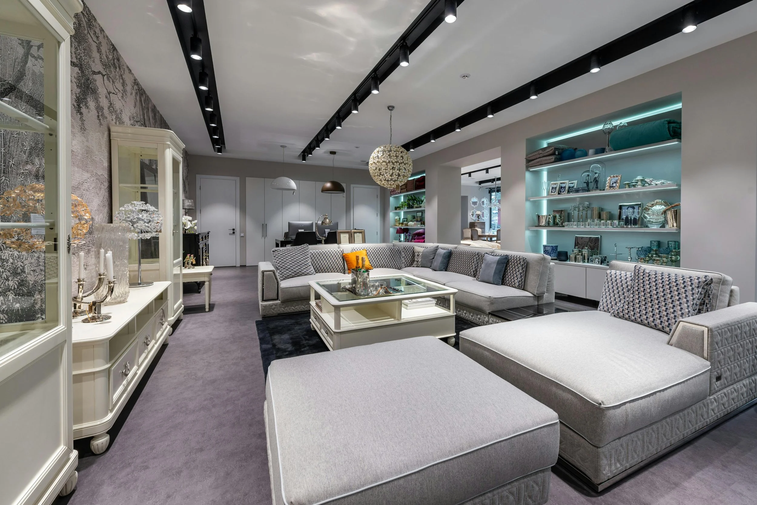A modern living room with a sectional sofa, glass coffee table, decorative shelving with lighting, and a textured accent wall.