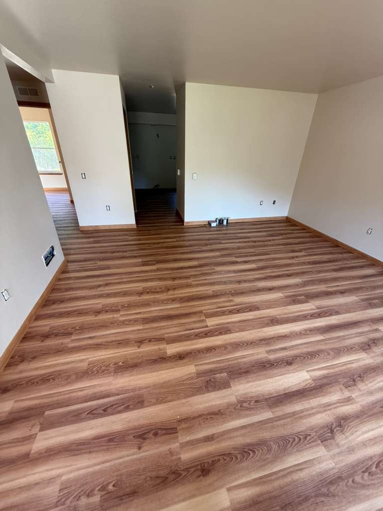 Empty room with new wood flooring, white walls, and a doorway leading to another room with similar floors. Multiple electrical outlets and vents are visible on the walls.