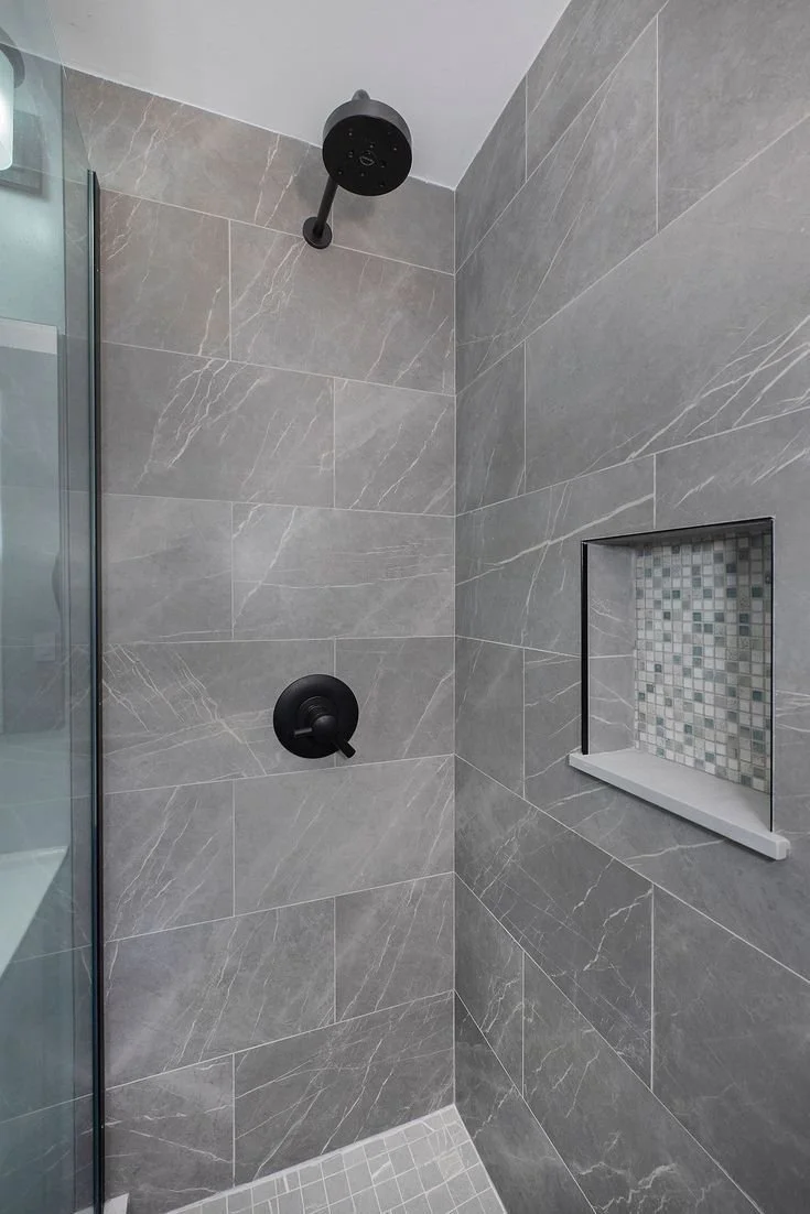 Gray tiled shower with black showerhead and control knob, built-in tiled shelf with mosaic tiles