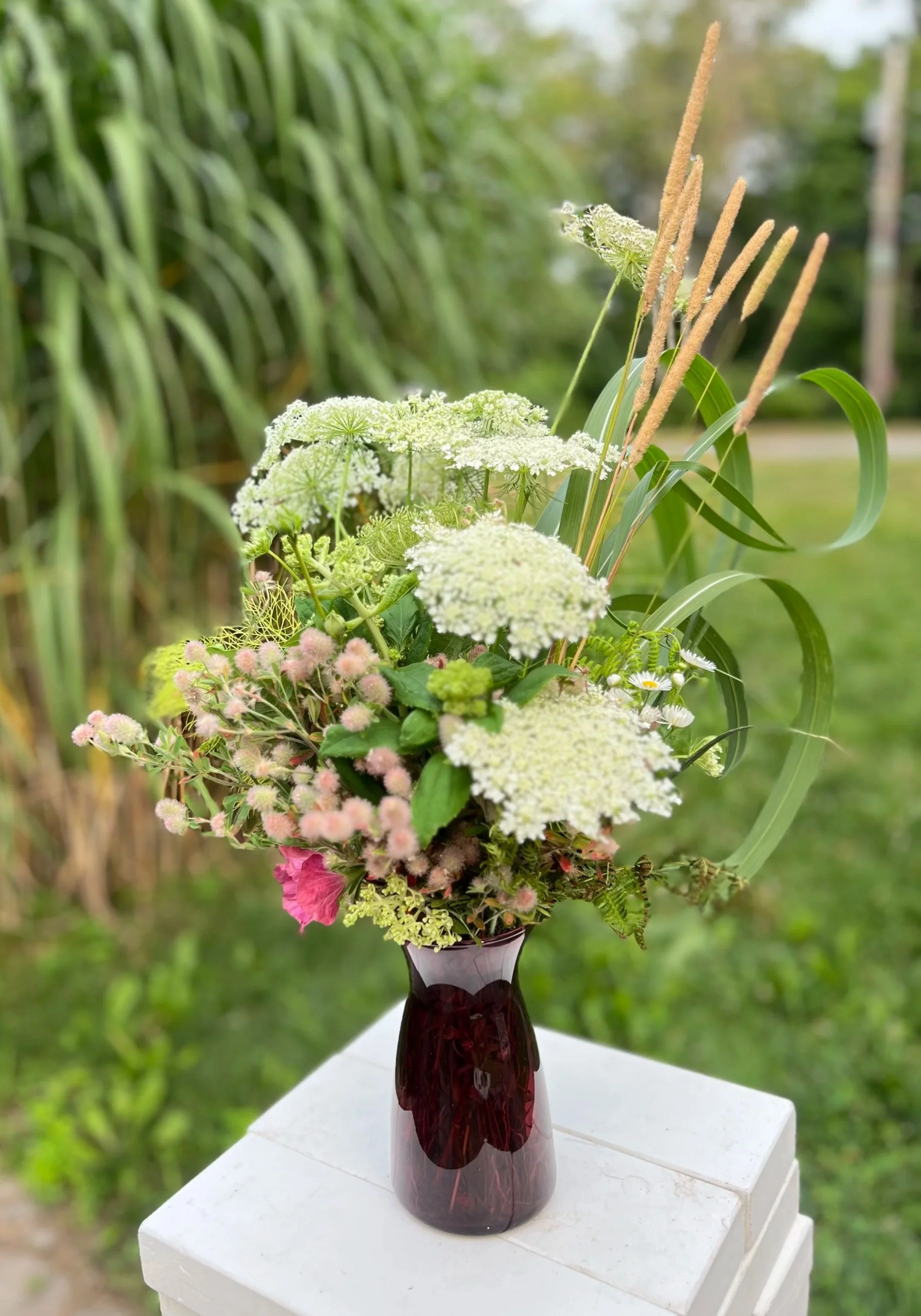 A bouquet of mixed flowers and greenery in a dark red glass vase placed on a white pedestal outdoors with green foliage in the background.