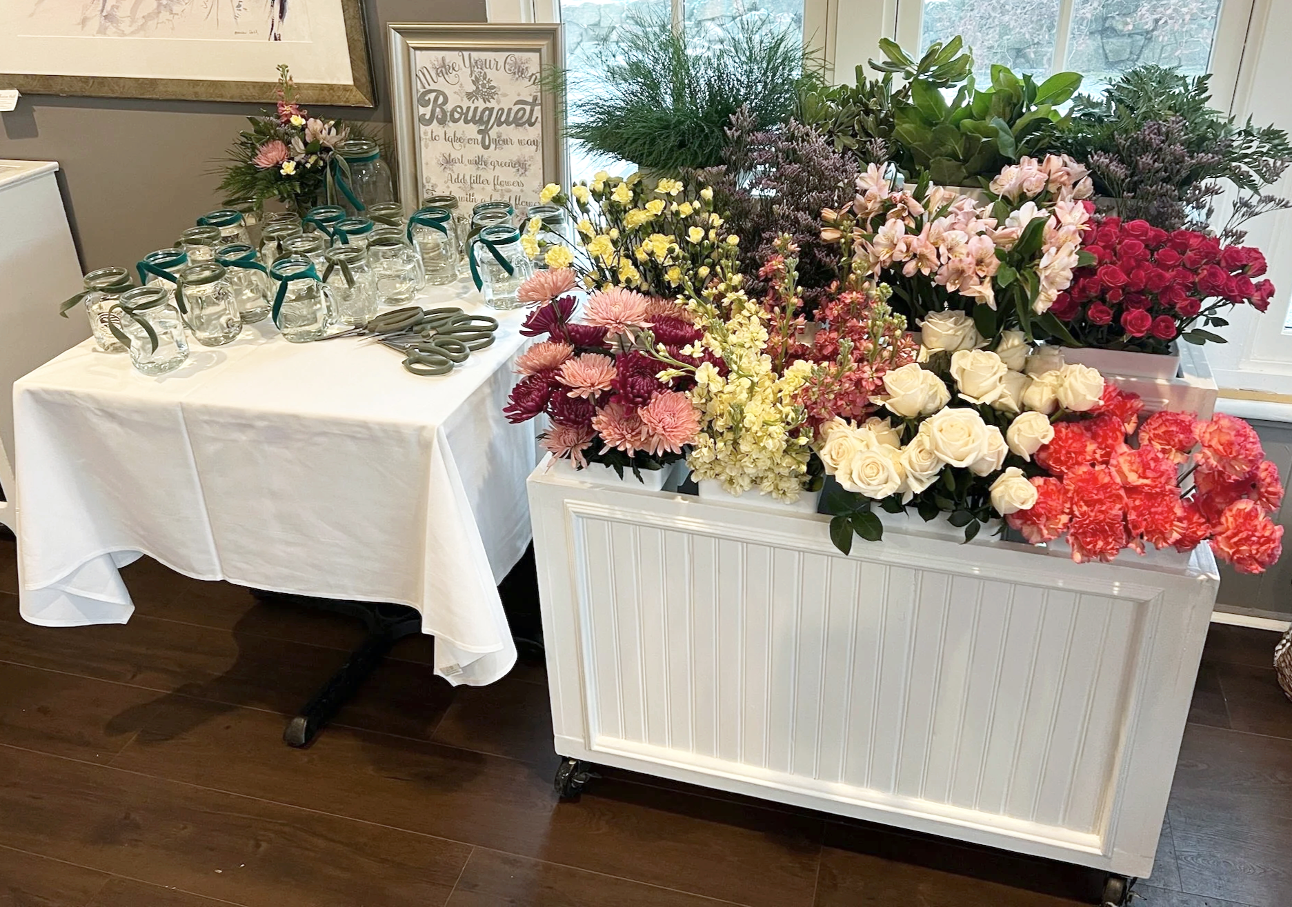 A table with a white tablecloth and scissors, and a large white wagon filled with various pink, red, yellow, white, and purple flowers, located indoors near a window.