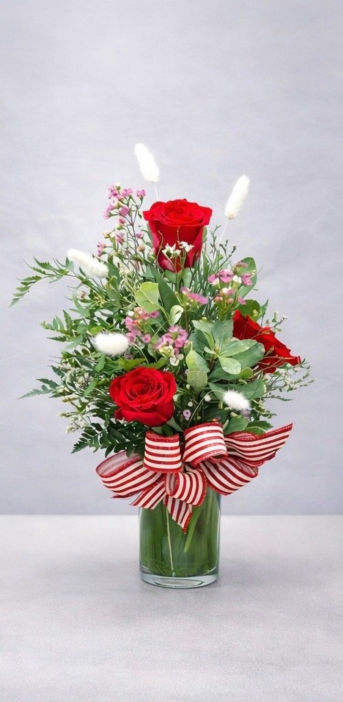 Red roses in a glass vase with green foliage, pink accents, white textured elements, and a red and white striped ribbon.