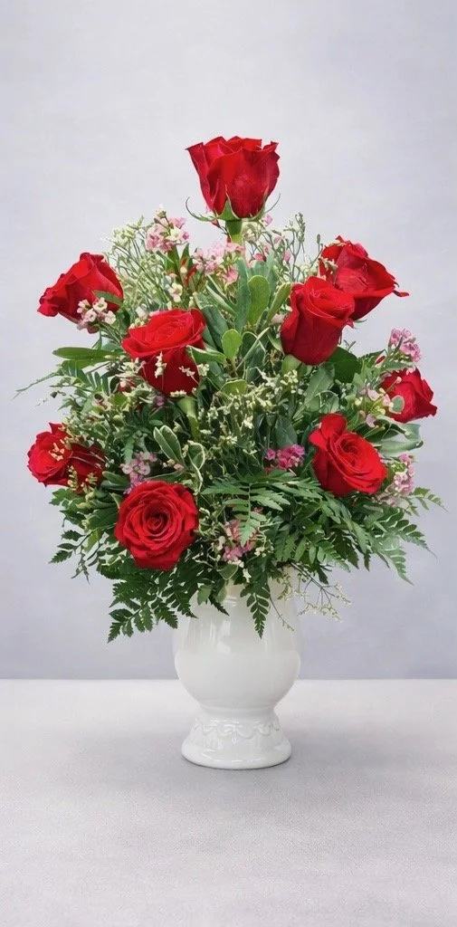 A bouquet of red roses and pink flowers in a white vase with green foliage, placed on a white surface against a light gray background.
