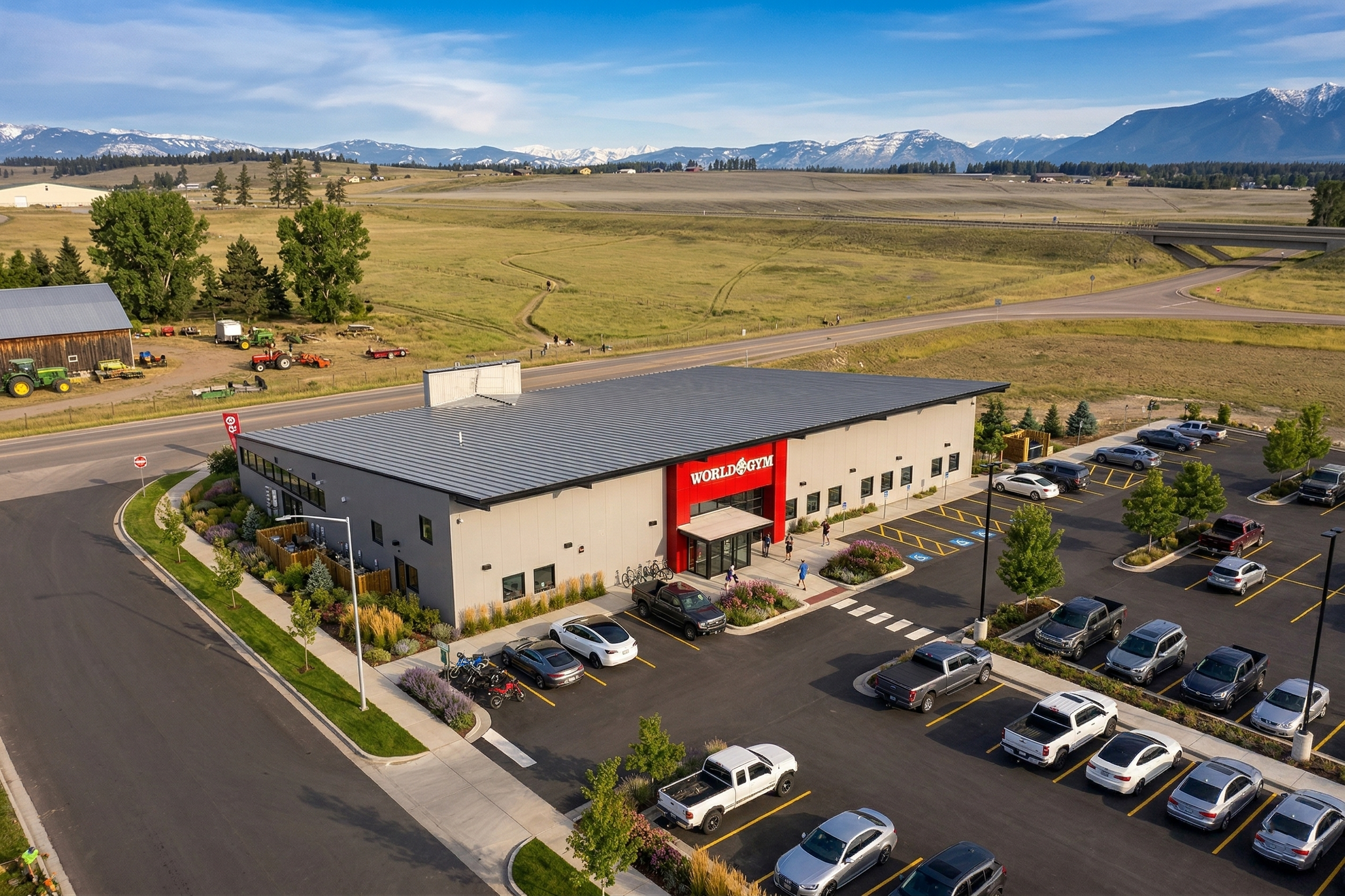 A large gym building with a gray exterior and a red sign that reads 'World Gym' on the front, surrounded by a parking lot with numerous parked cars, trees, and landscaped areas. In the background, there are open fields, trees, and snow-capped mountai