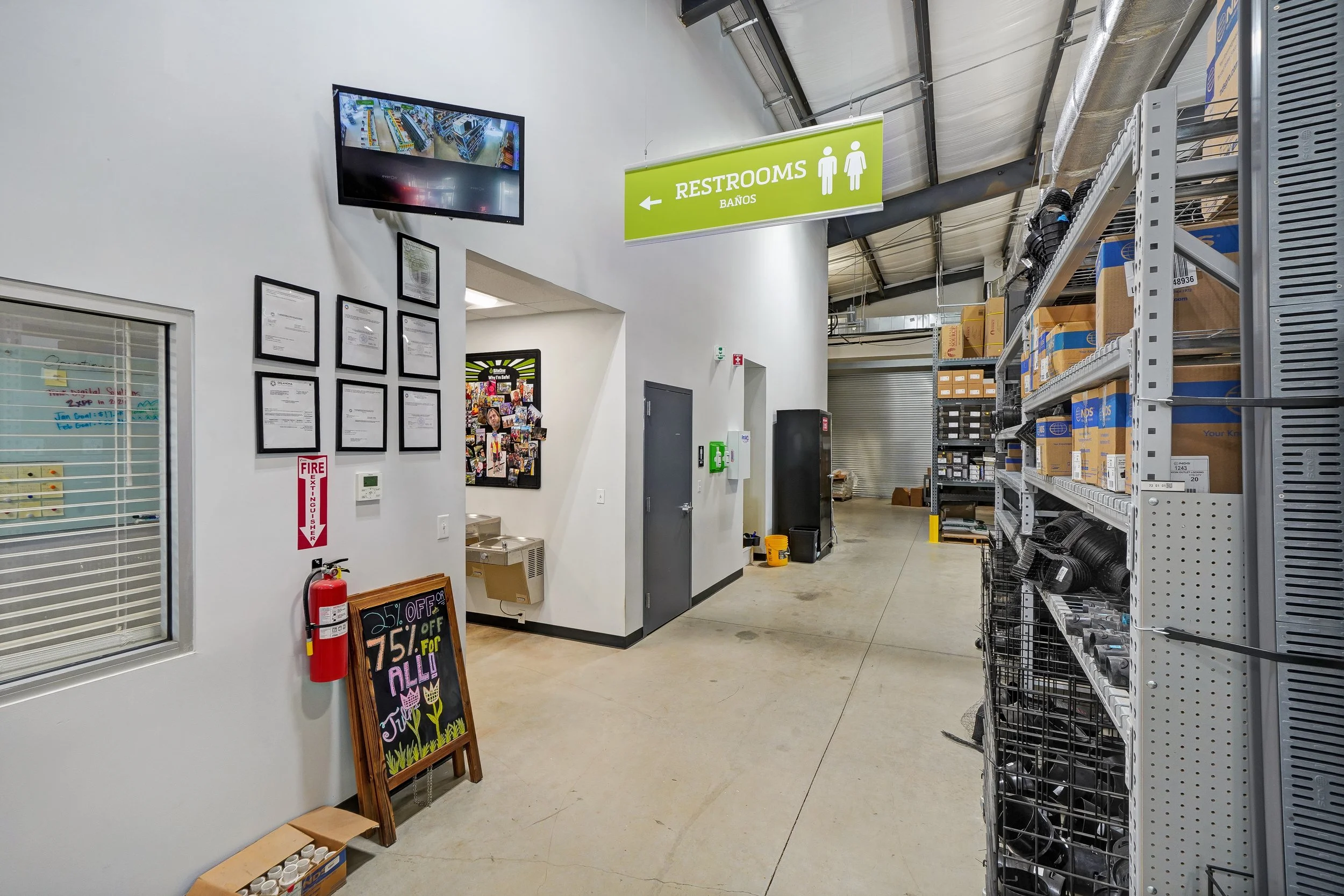 Interior of a warehouse or store with a sign pointing to restrooms, shelving with boxes and equipment, a fire extinguisher, and a chalkboard sign advertising discounts.